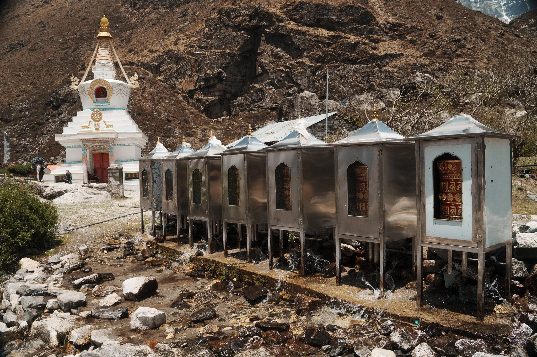 A small white Buddhist stupa with golden decorations and a series of metallic water fountains in front, set against rocky mountain terrain.