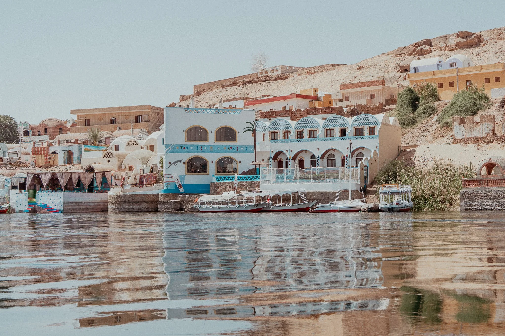 Colorful buildings with blue accents on a hillside next to a body of water, with boats docked along the shore.