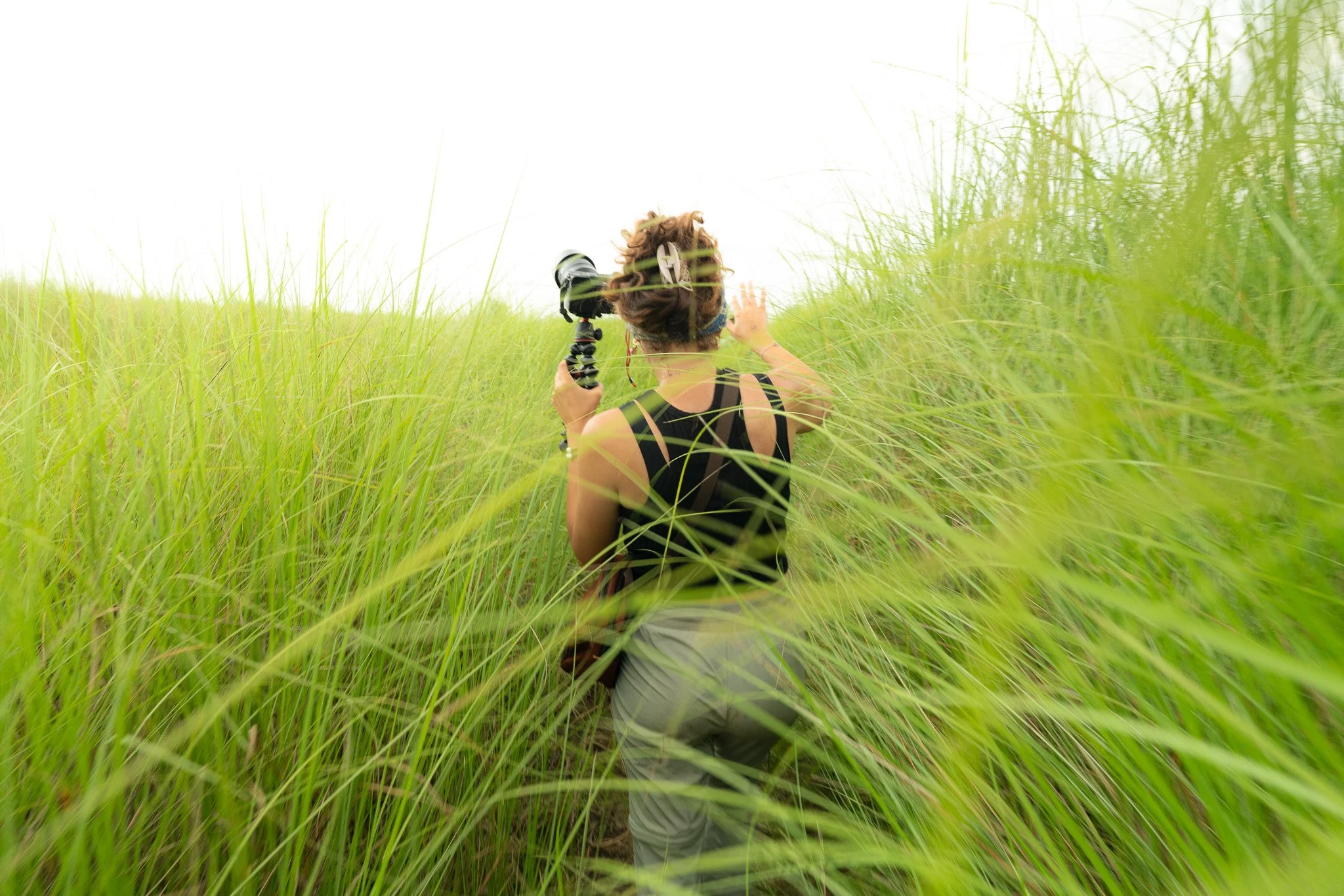 A person with short hair and a black tank top kneeling in tall green grass, holding a camera, with their back to the camera, amidst a lush field.