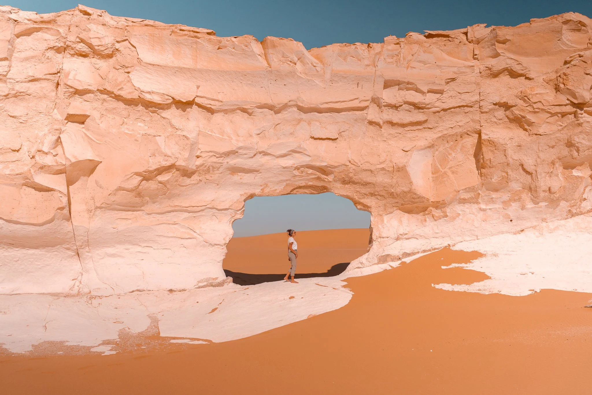 A person standing in front of a large desert rock formation with a natural arch, surrounded by sand dunes and a blue sky.