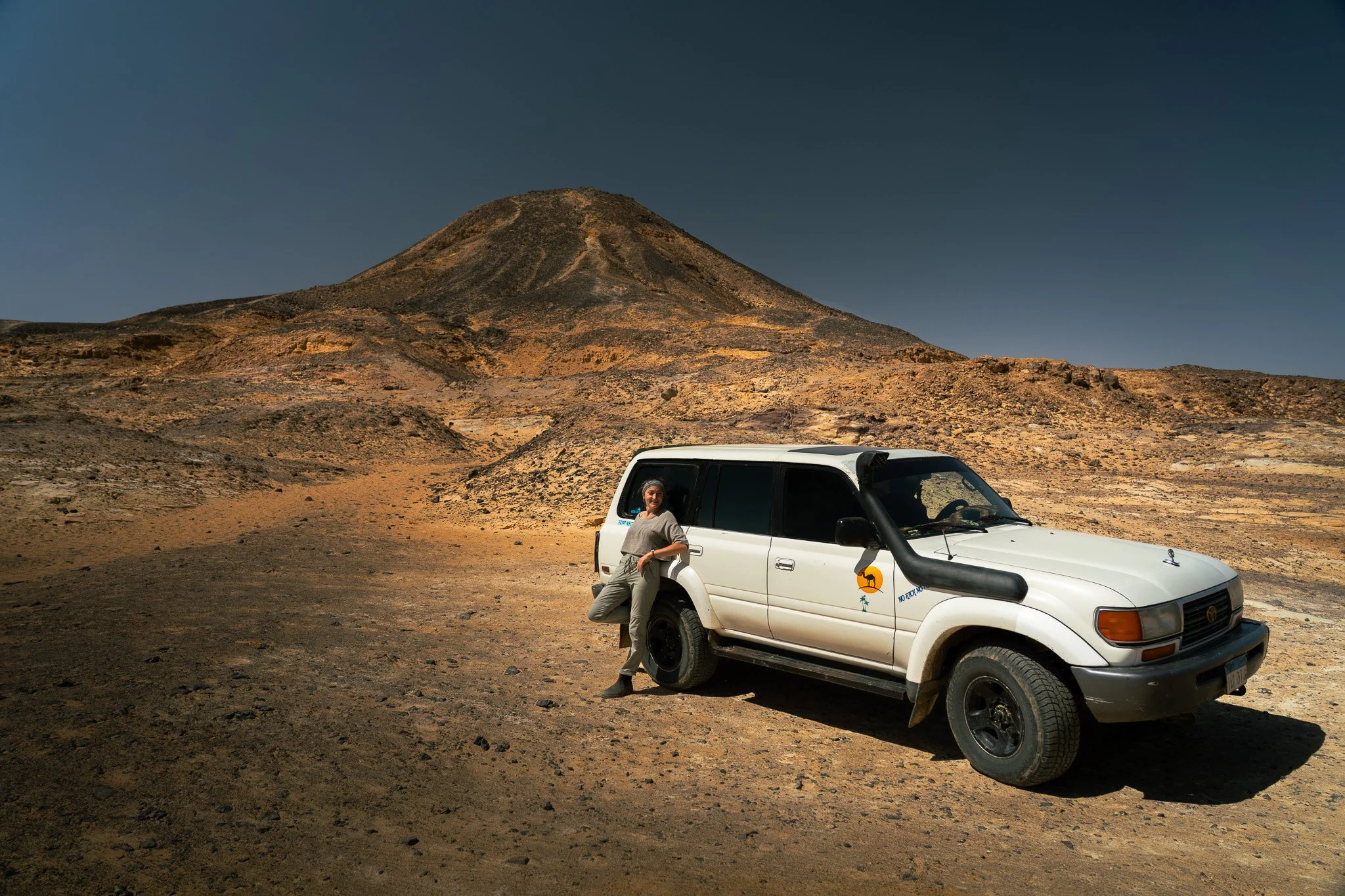 A woman in outdoor clothing leaning against a white SUV in a rocky desert landscape with a mountain in the background, under a clear sky.