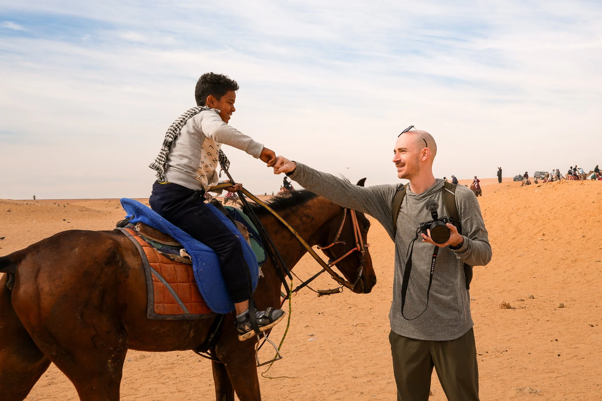 A young boy riding a horse in a desert, fist bumping with an adult man holding a camera, with other people and vehicles in the background.