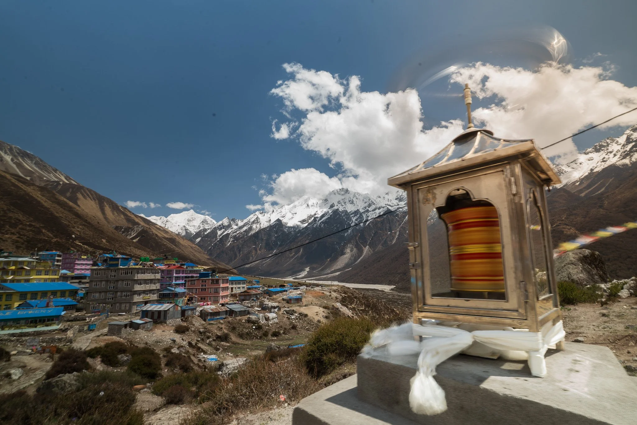 A prayer wheel in a metal enclosure with a mountain village and snow-capped peaks in the background.
