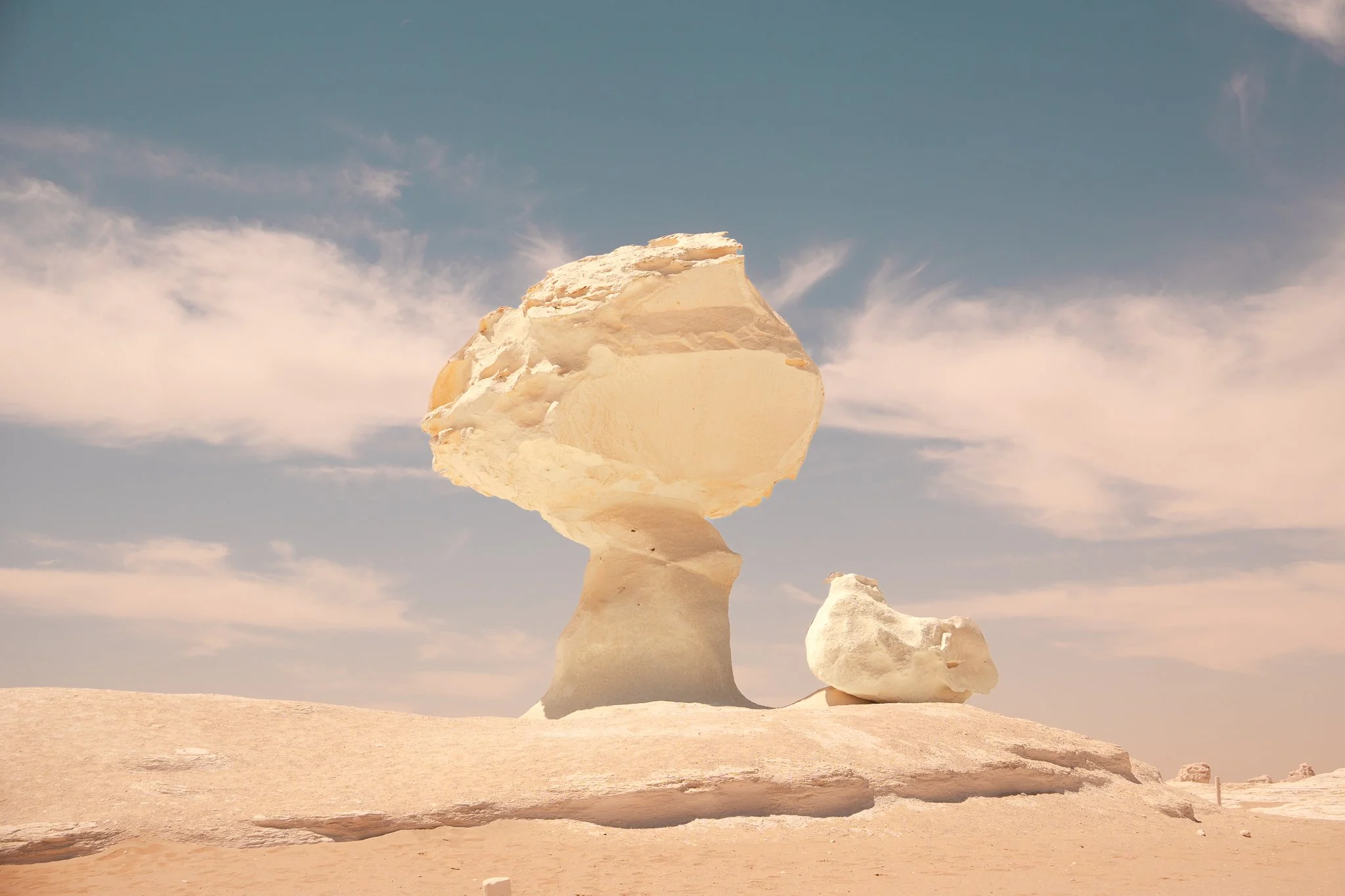 Large white rock formation resembling a mushroom in a desert landscape with a blue sky and scattered clouds