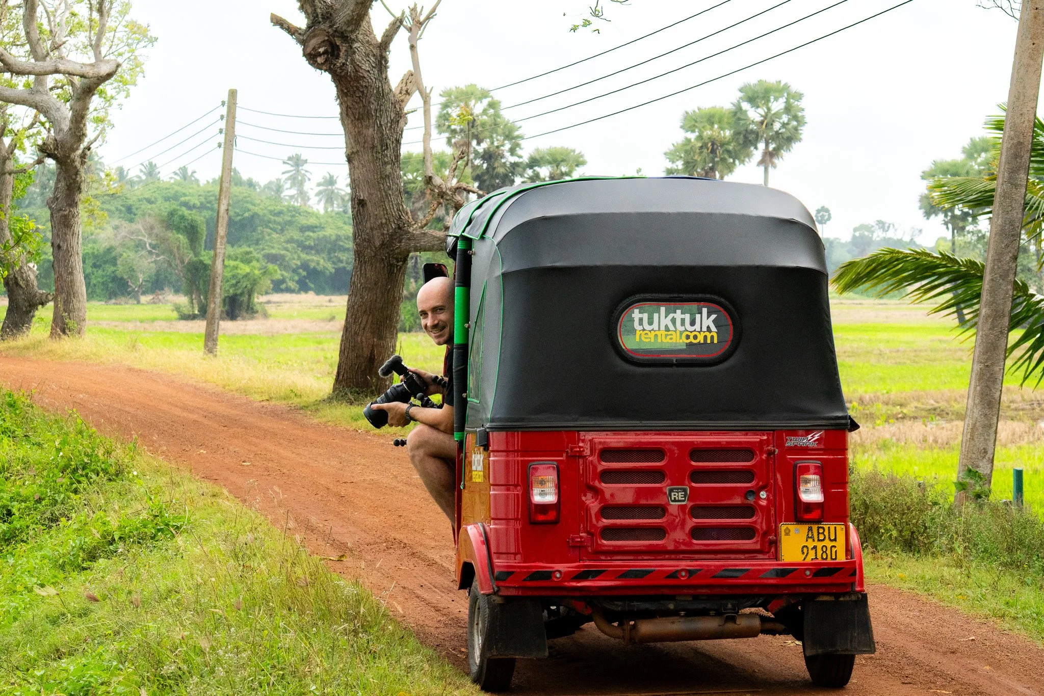A person peeking out from the back of a red tuk-tuk vehicle, holding a camera, on a dirt road surrounded by trees and greenery.