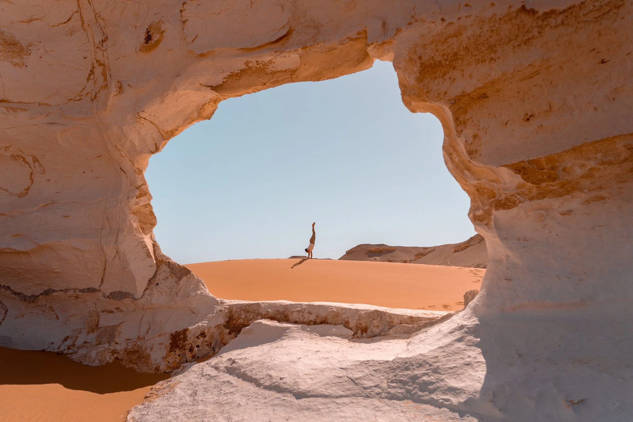 Person performing a handstand on sandy desert dunes, framed by a white rock formation with a sky in the background.