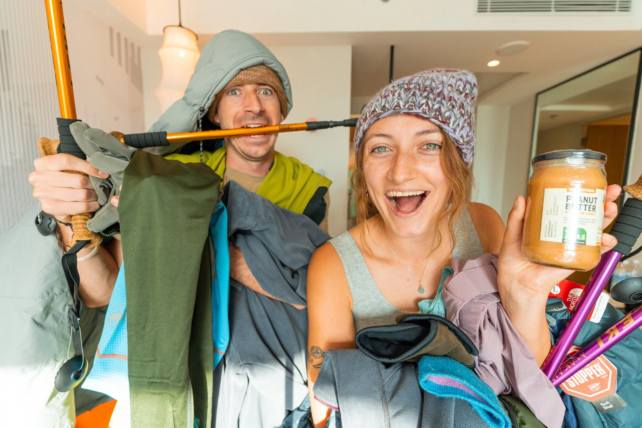 Two smiling hikers, one woman and one man, holding gear and a jar of peanut butter, indoors.