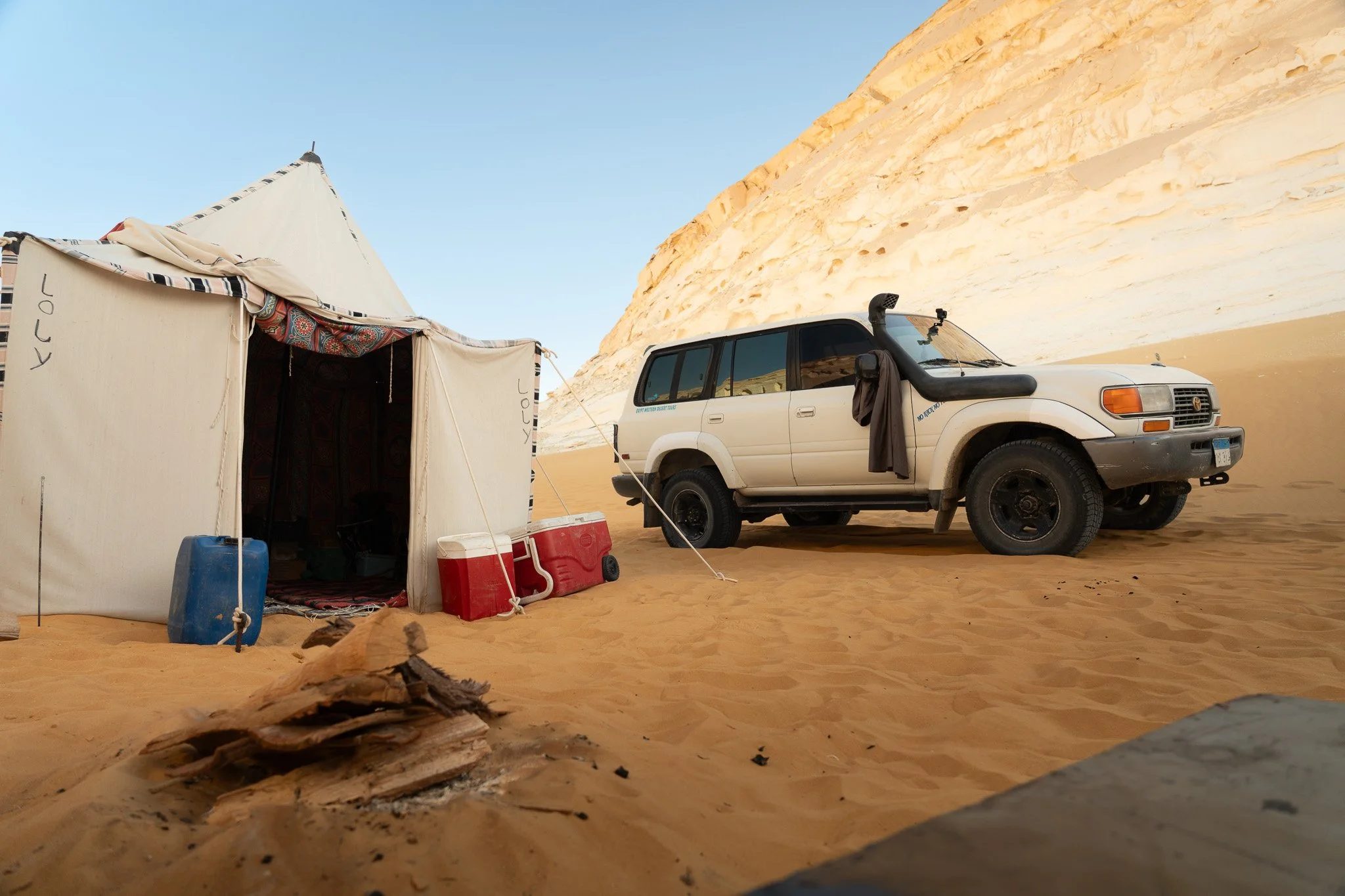 A camping scene in a desert with a beige tent, water coolers, and a white SUV parked on sandy terrain near a large rock formation under a clear sky.