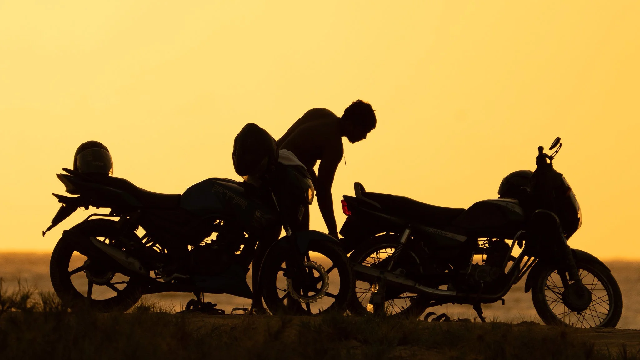 Silhouette of a person leaning over two motorcycles during sunset or sunrise.