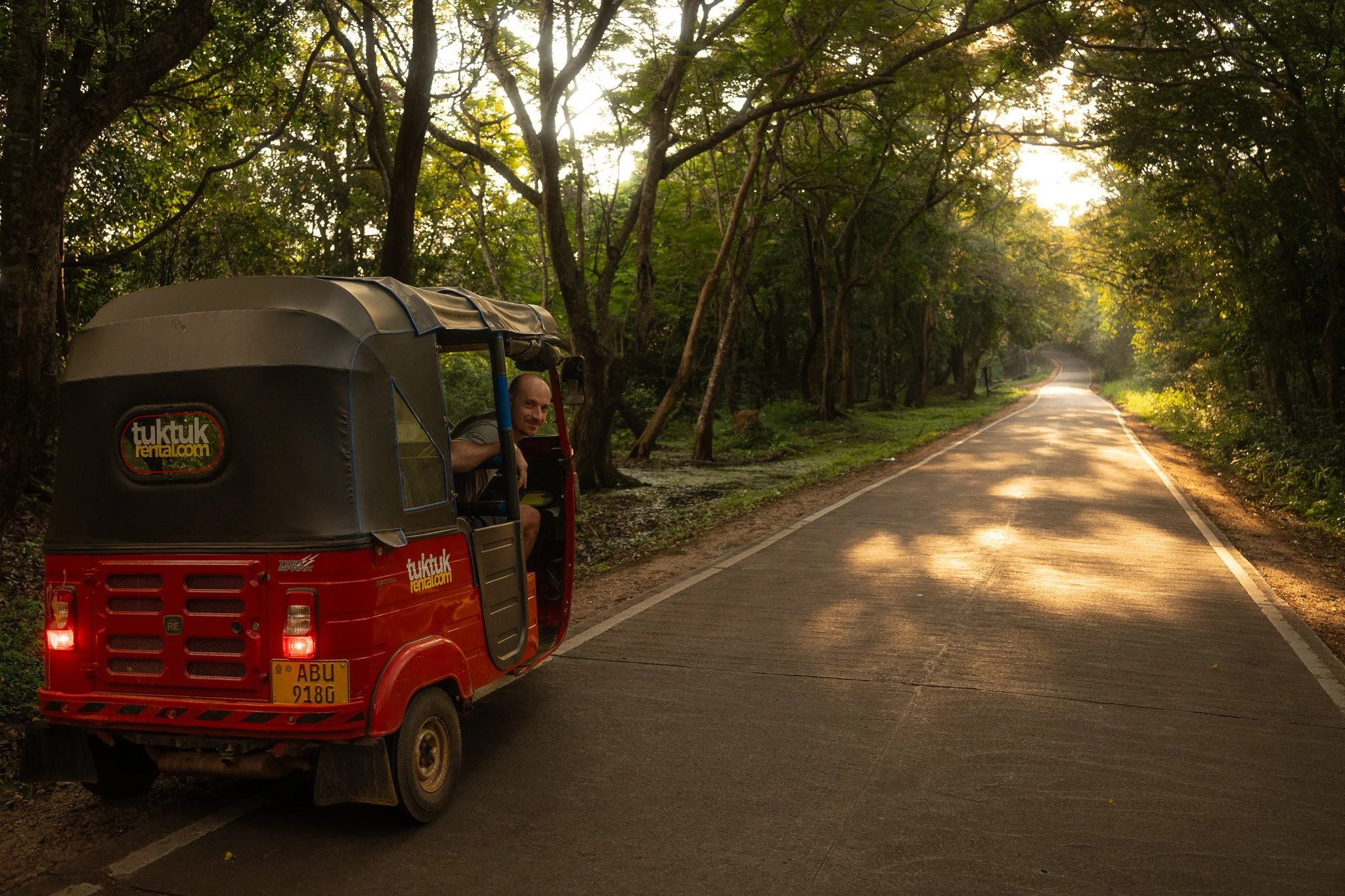 A man in a tuk-tuk vehicle, parked on a rural road surrounded by trees during sunset.