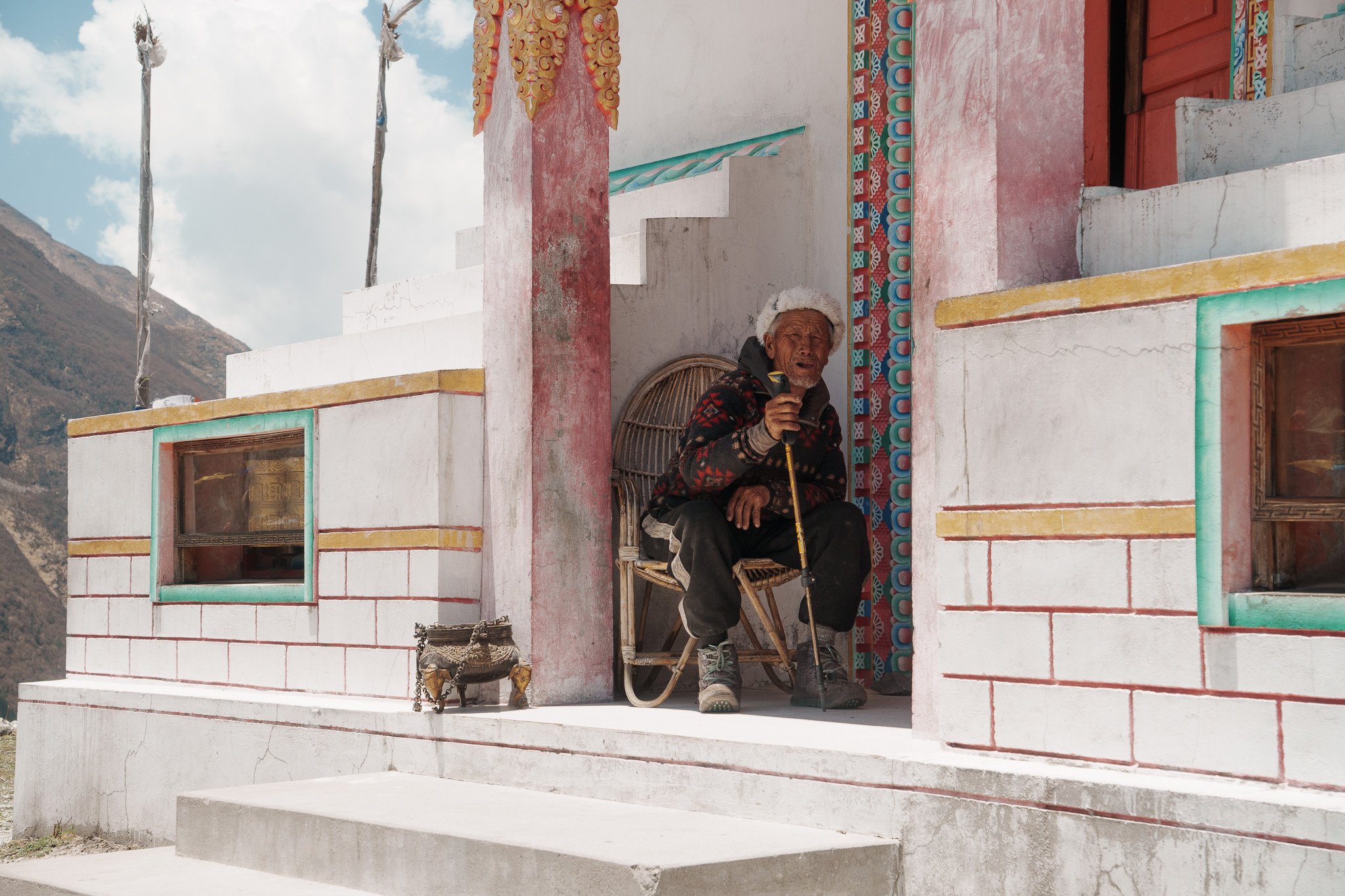 An elderly man sitting on a chair outside a colorful temple, holding a walking cane and wearing a hat, with a mountain in the background.