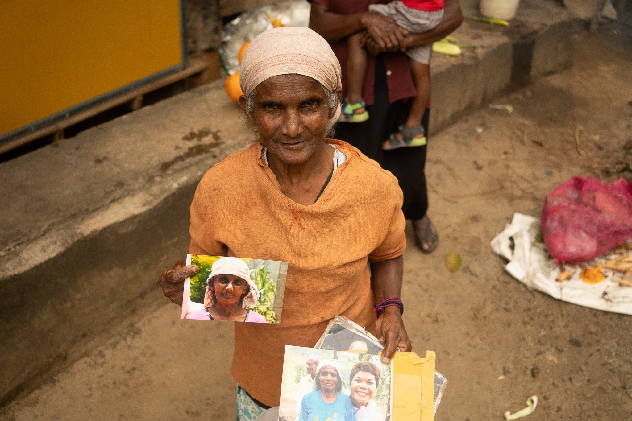 An elderly woman with gray hair under a beige headscarf holding photographs of herself in different stages of life. She is dressed in an orange top and standing on a dirt street.