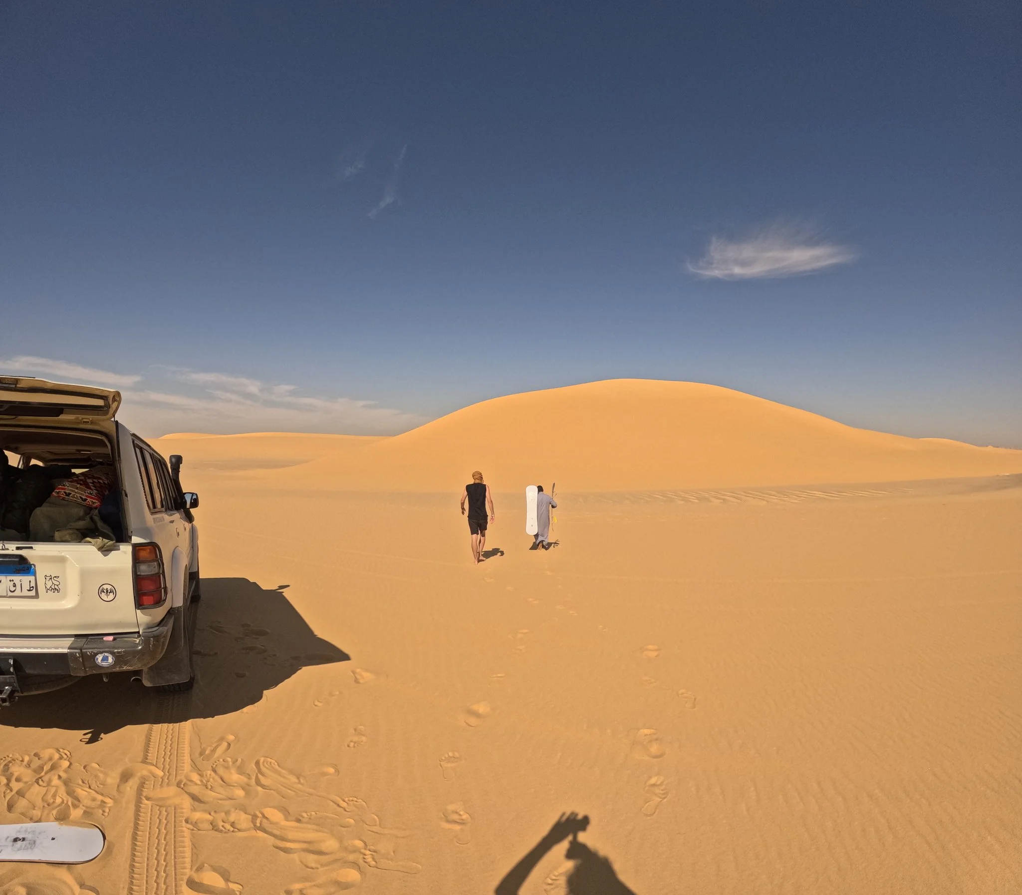 Sand dunes in a desert with two people walking towards the horizon, one carrying a snowboard, and an open vehicle on the left side, under a blue sky with a few clouds.