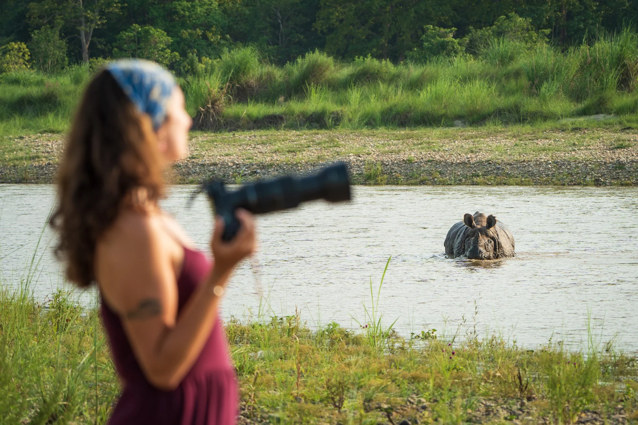 A woman standing near a river holding a camera, with a rhinoceros in the water in the background.