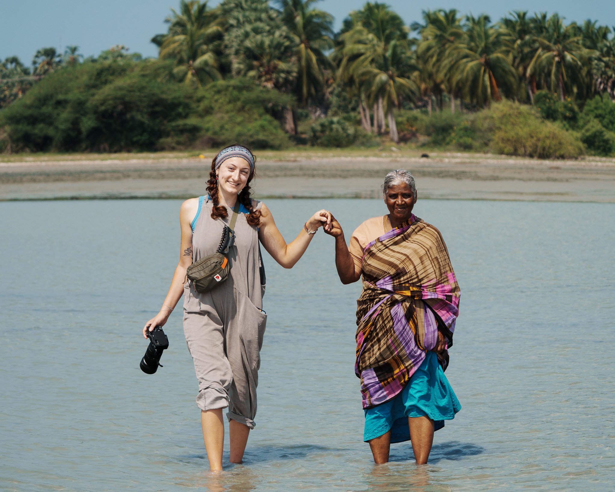 A young woman and an older woman holding hands while standing in shallow water on a beach with palm trees in the background.