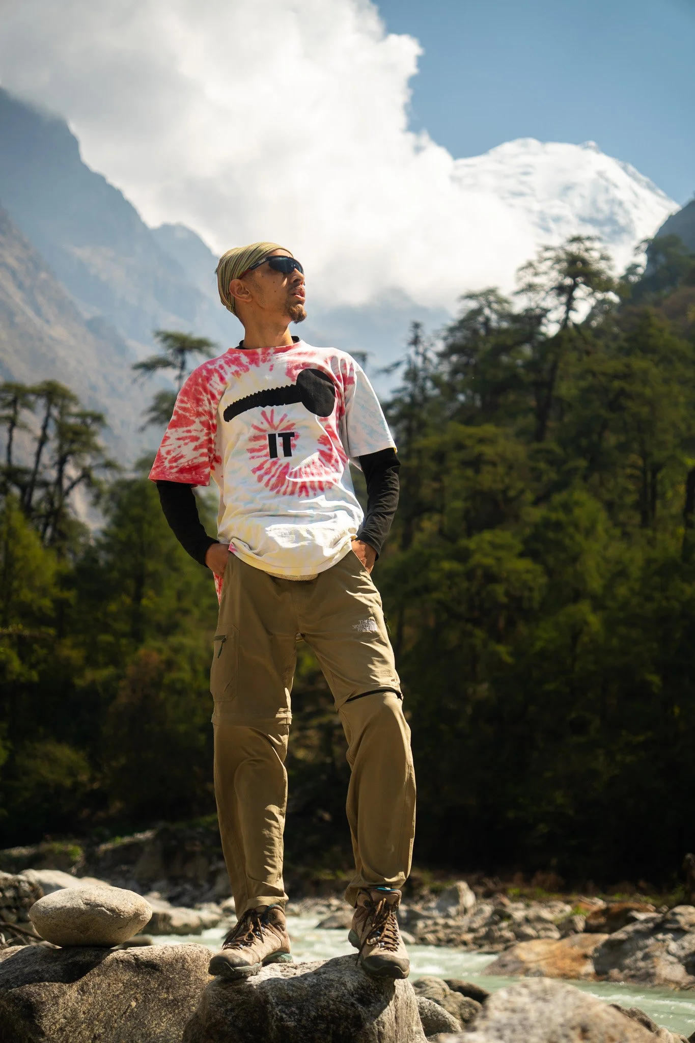 A man standing outdoors on rocky terrain with a mountain landscape in the background, wearing sunglasses, a tie-dye t-shirt with the words 'I ❤️ IT', long-sleeve undershirt, cargo pants, and hiking boots.