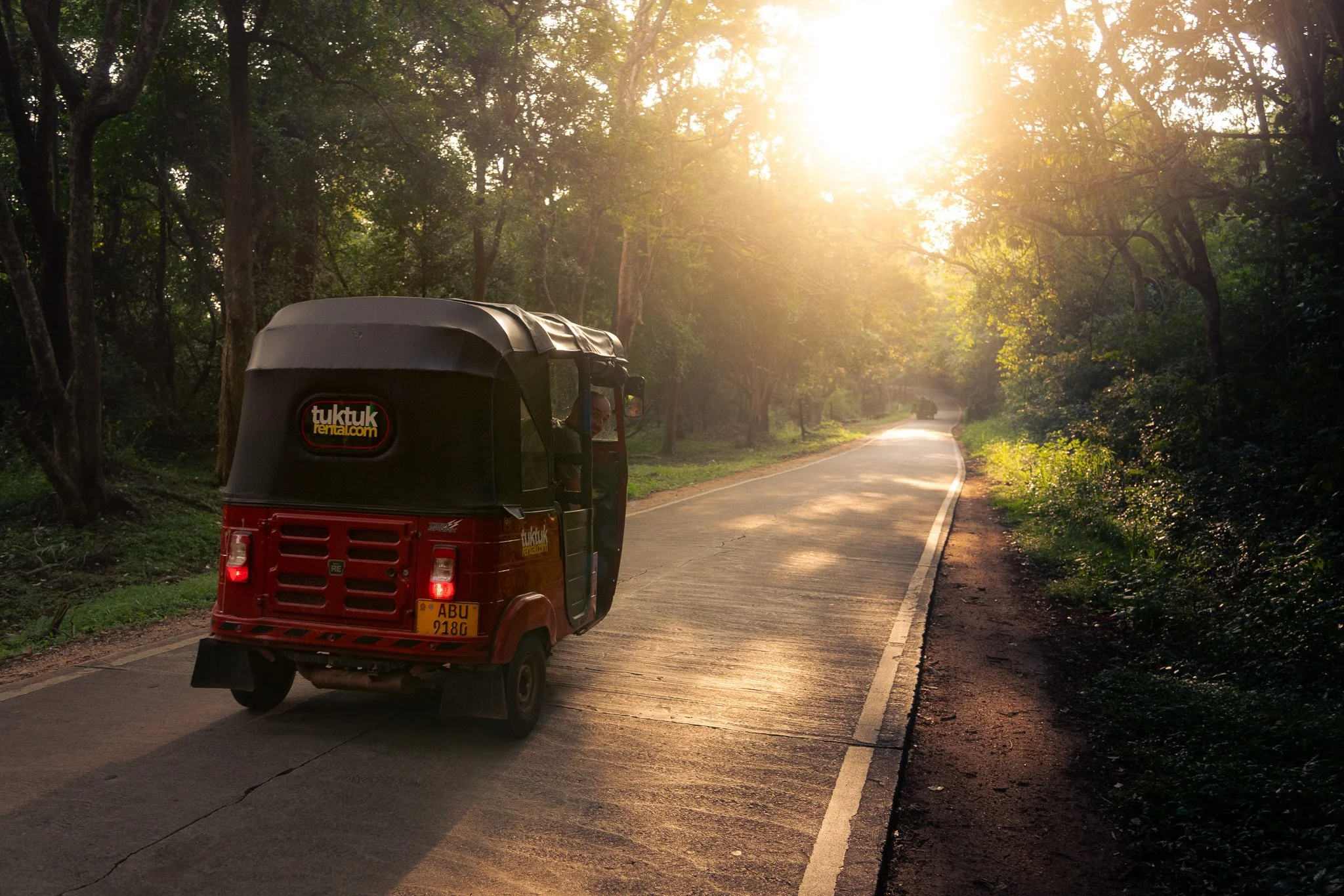 A red tuk-tuk on a winding road with trees and foliage on both sides, sunlight shining through the trees during sunset or sunrise.