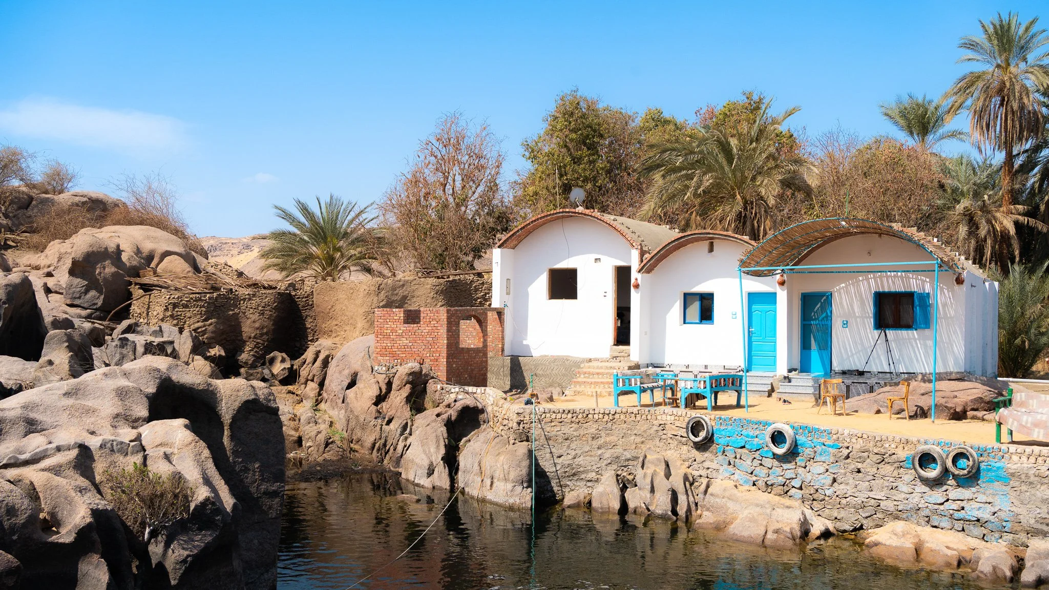 Coastal house with white walls, blue doors, and windows, surrounded by palm trees and rocks, next to a body of water under a clear blue sky.