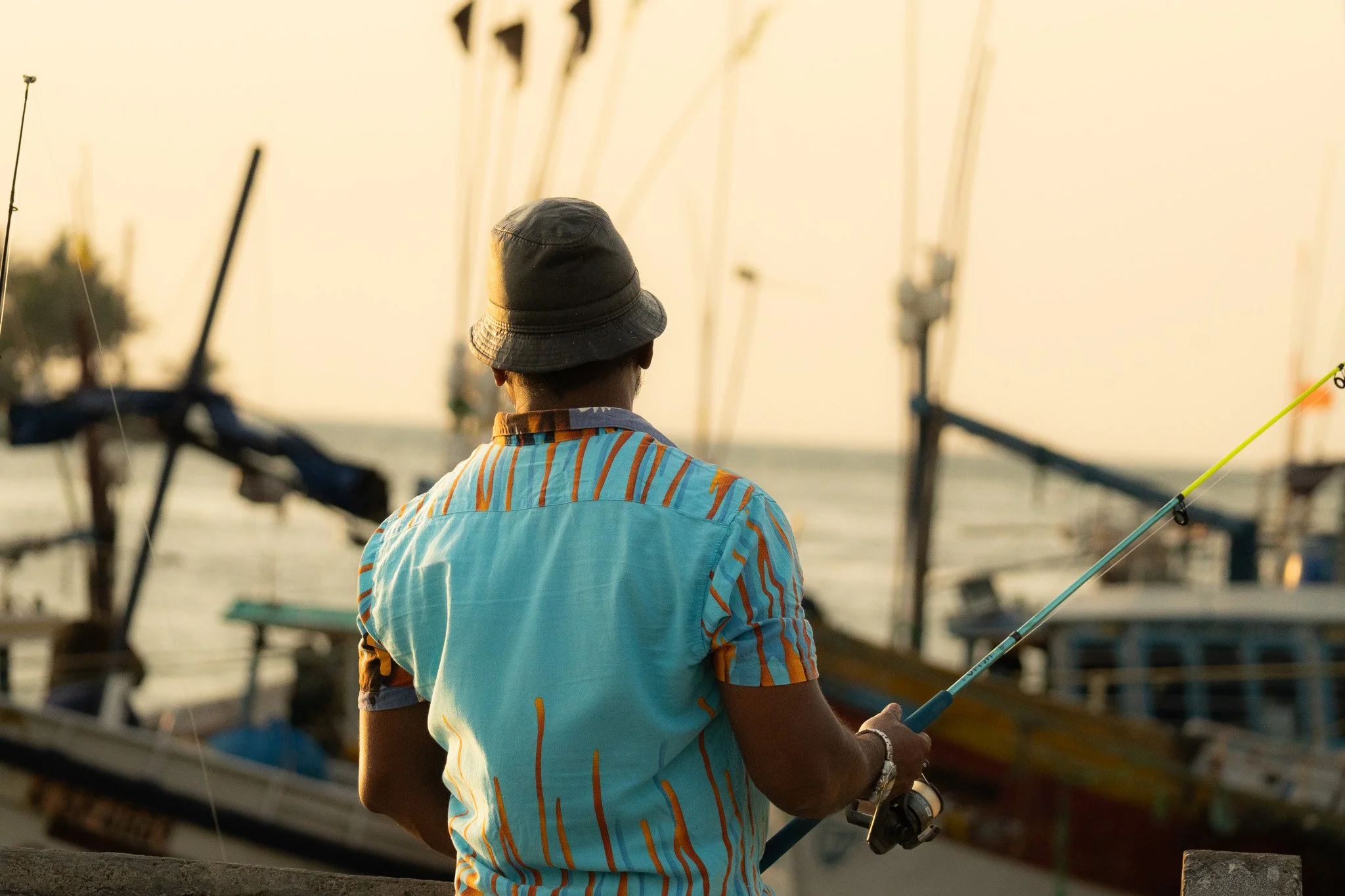 A person wearing a black hat and a colorful striped shirt, fishing near a boat dock at sunset.