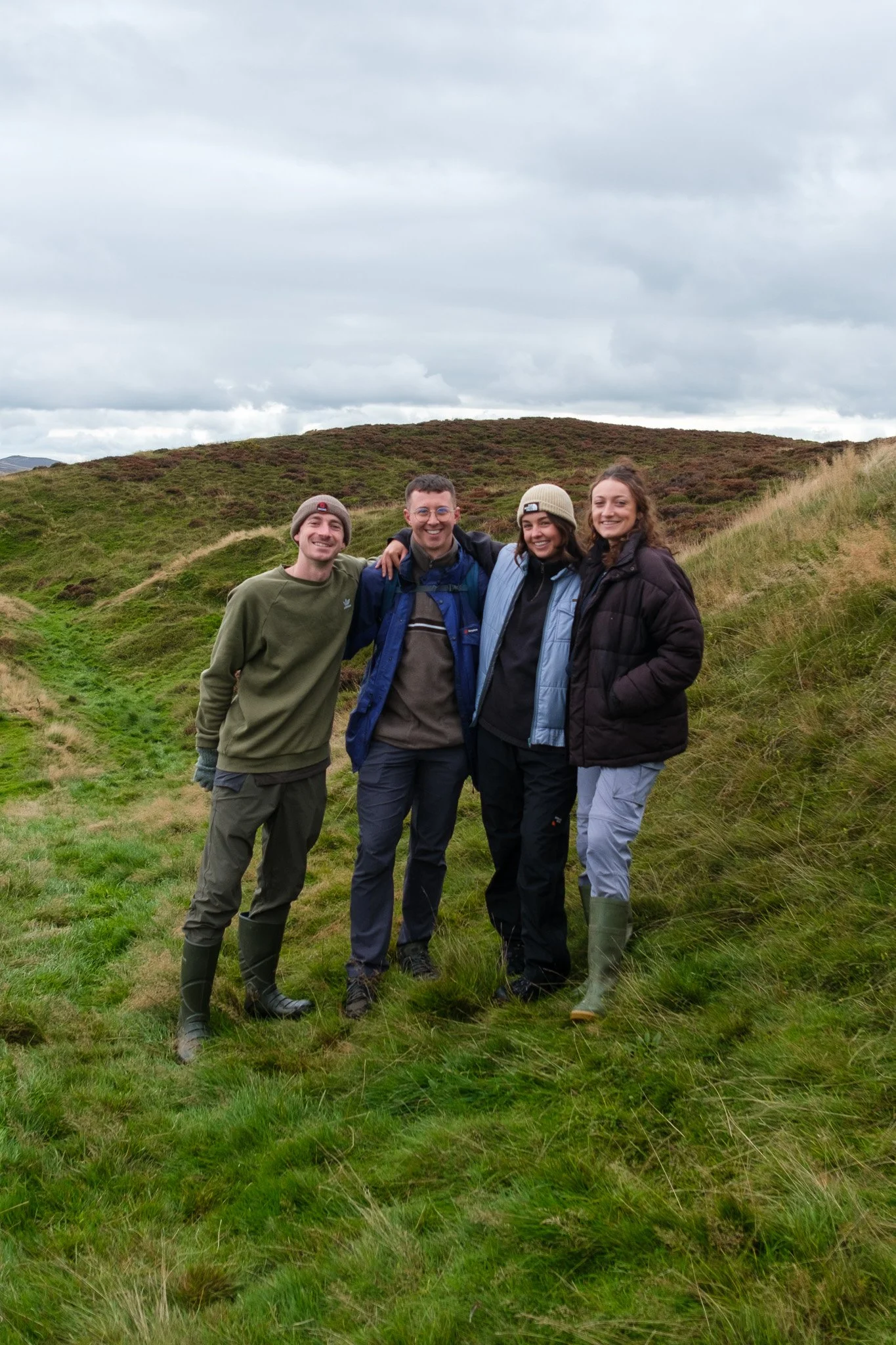 Four people standing on a grassy hillside with cloudy sky, dressed in outdoor clothing and hiking gear.
