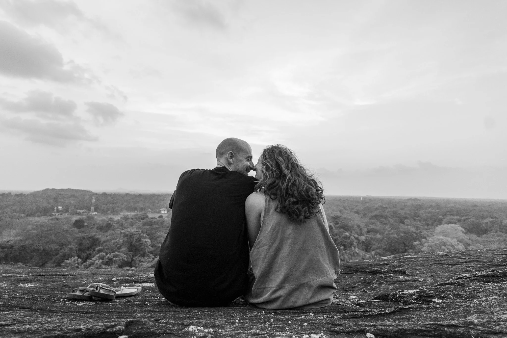 A black-and-white photo of a couple sitting on a rocky ledge, facing each other and smiling, with a scenic landscape of trees and a cloudy sky in the background.