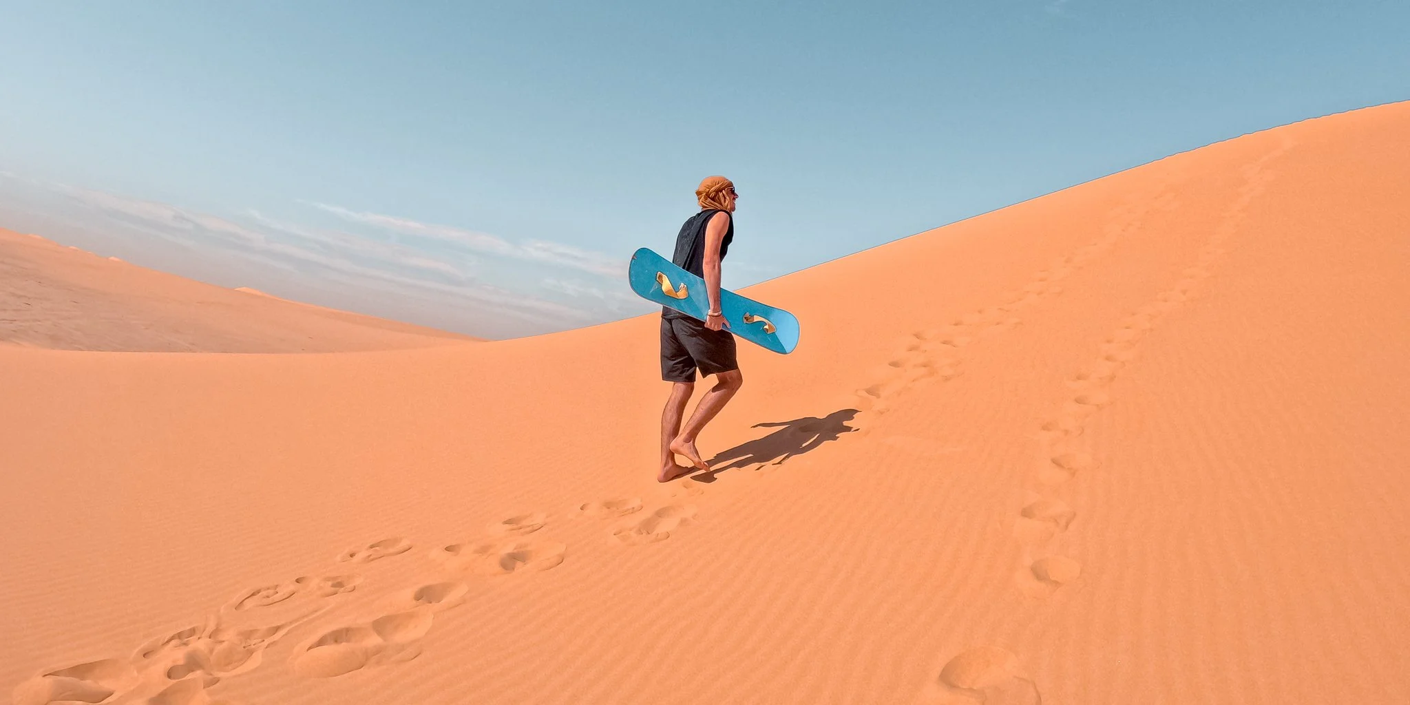 A man in black shorts and a sleeveless top carrying a blue skateboard walks up sand dunes in a desert under a partly cloudy sky.