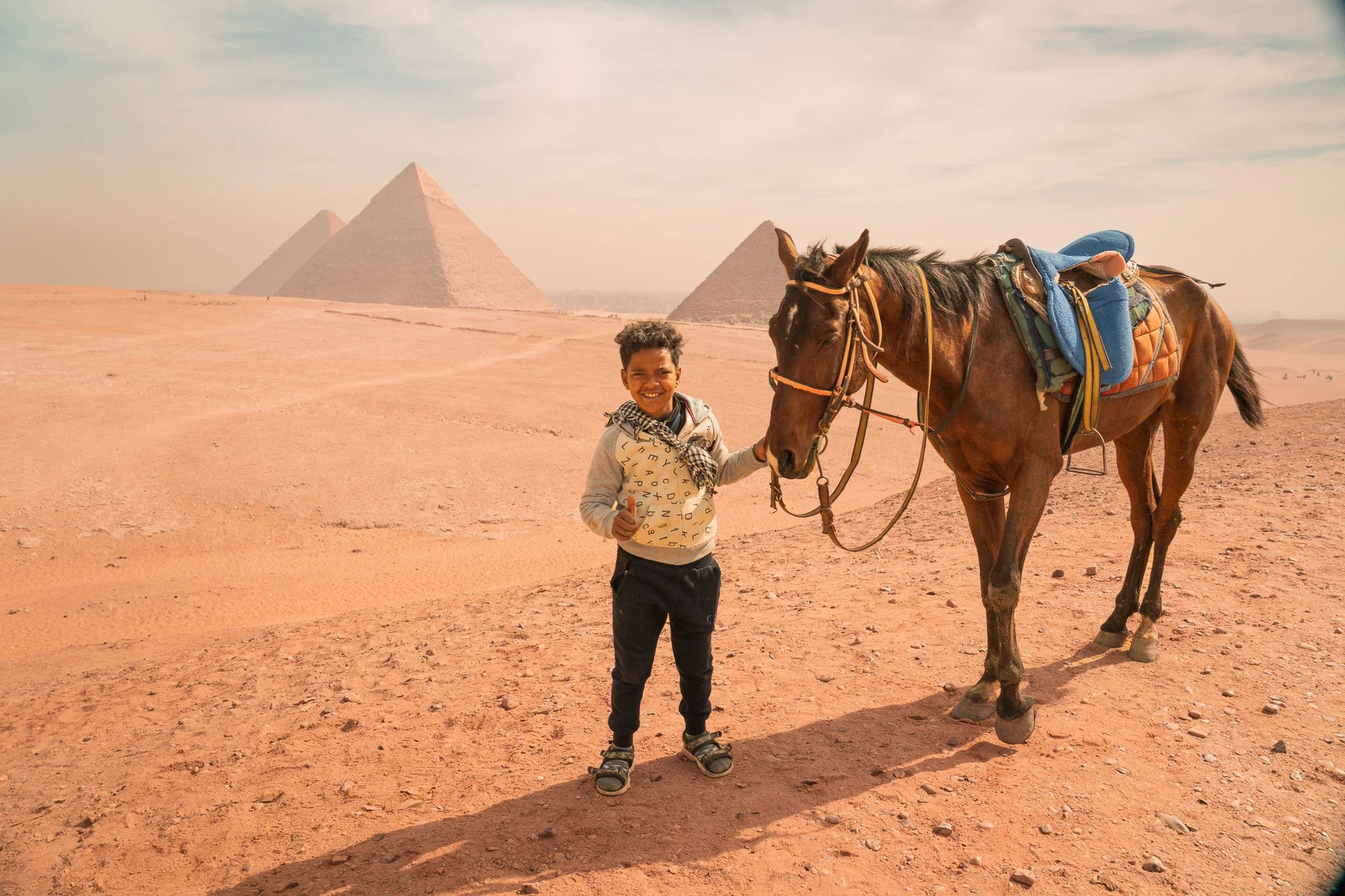 A young boy smiling and giving a thumbs up while holding a horse's bridle in the desert near the Pyramids of Giza, Egypt, during daytime.