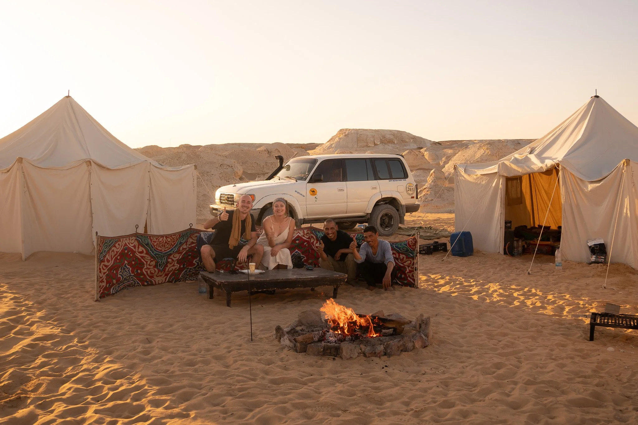 Group of five people sitting around a campfire in a desert with tents and a white off-road vehicle in the background.