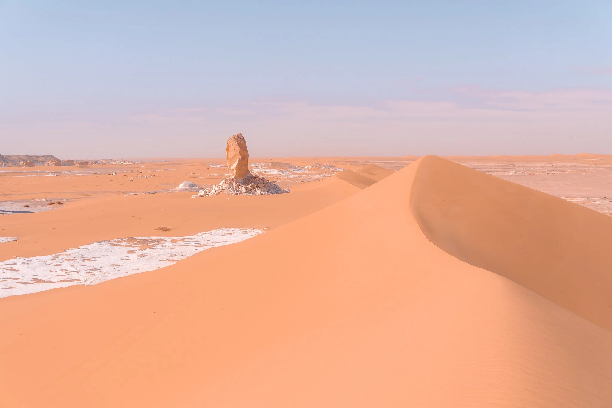 A vast desert landscape with sand dunes, a rock formation in the distance, and a clear blue sky.