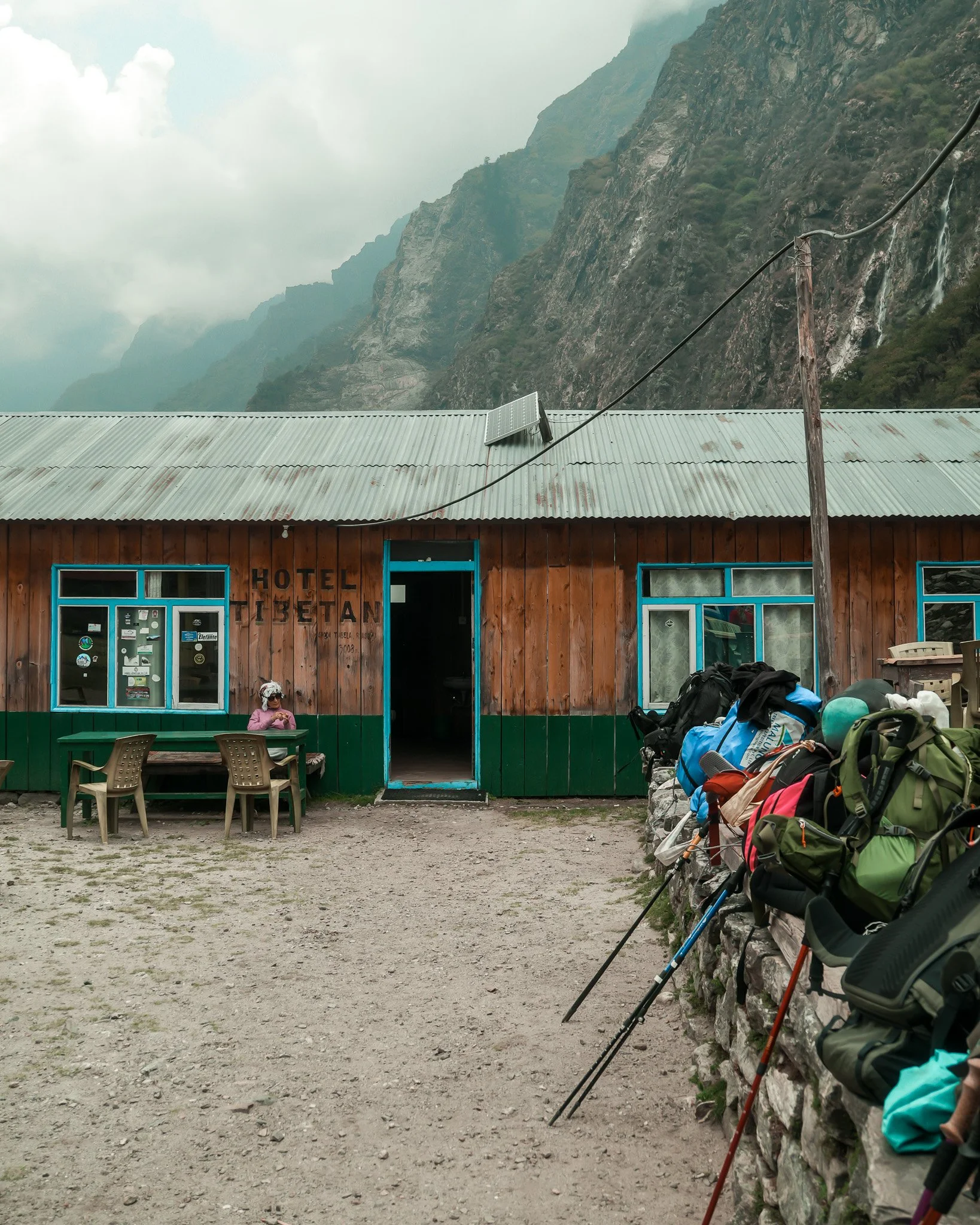 A wooden building with a sign that reads 'Hotel Isetan,' with mountains in the background, backpacks and hiking gear lined up outside, and a person sitting at a table.
