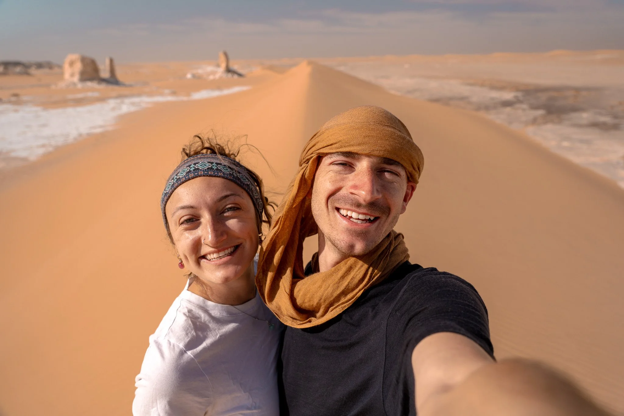 A smiling couple taking a selfie on a sand dune in a desert, with a blue sky and distant rock formations in the background.