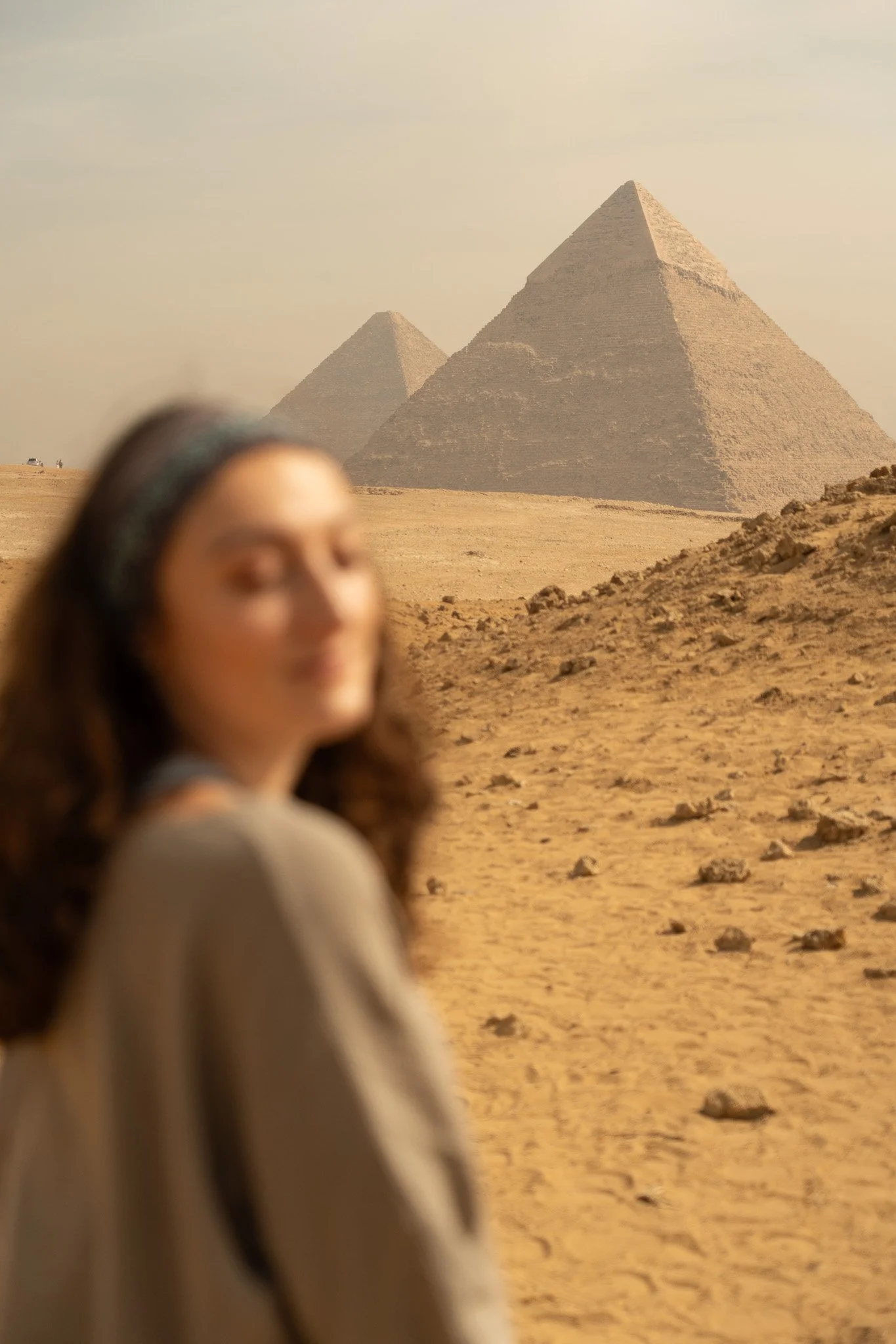 A woman with long hair and a headband standing in the desert with the pyramids of Giza in the background.