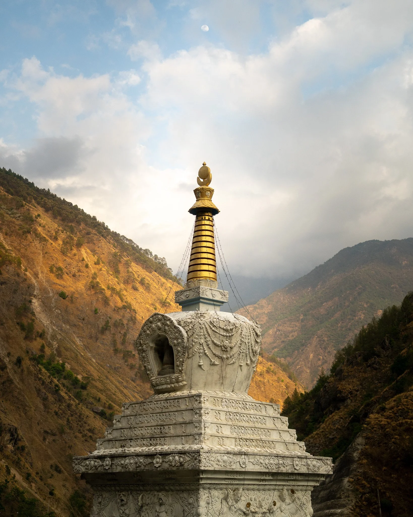A white stupa with intricate carvings and a golden spire, set against a mountainous landscape with cloudy skies.