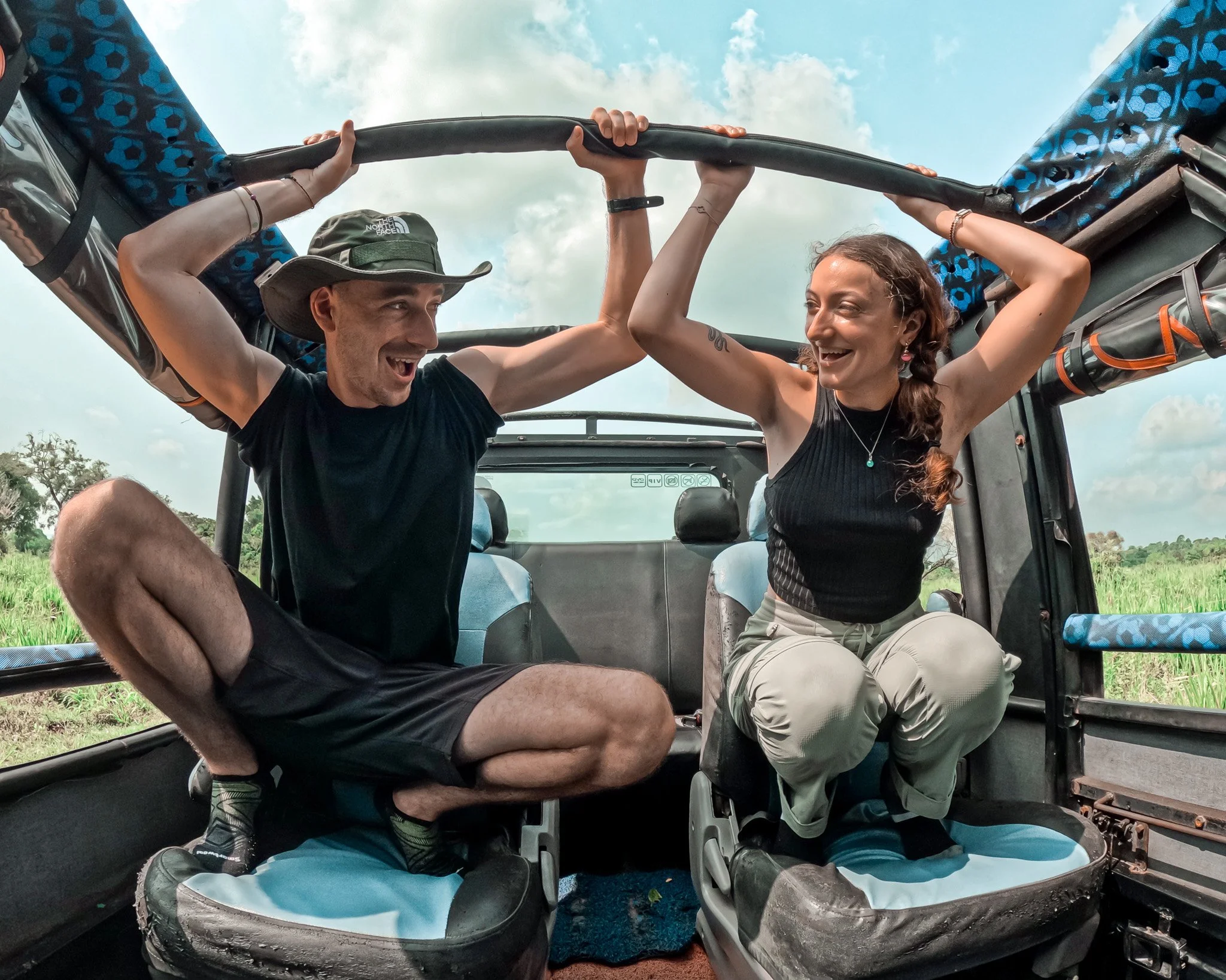 Two people, a man and a woman, sitting inside a vehicle. They are smiling, holding the roof of the vehicle with their hands, and appear to be excited. The background shows a grassy field and a cloudy sky.