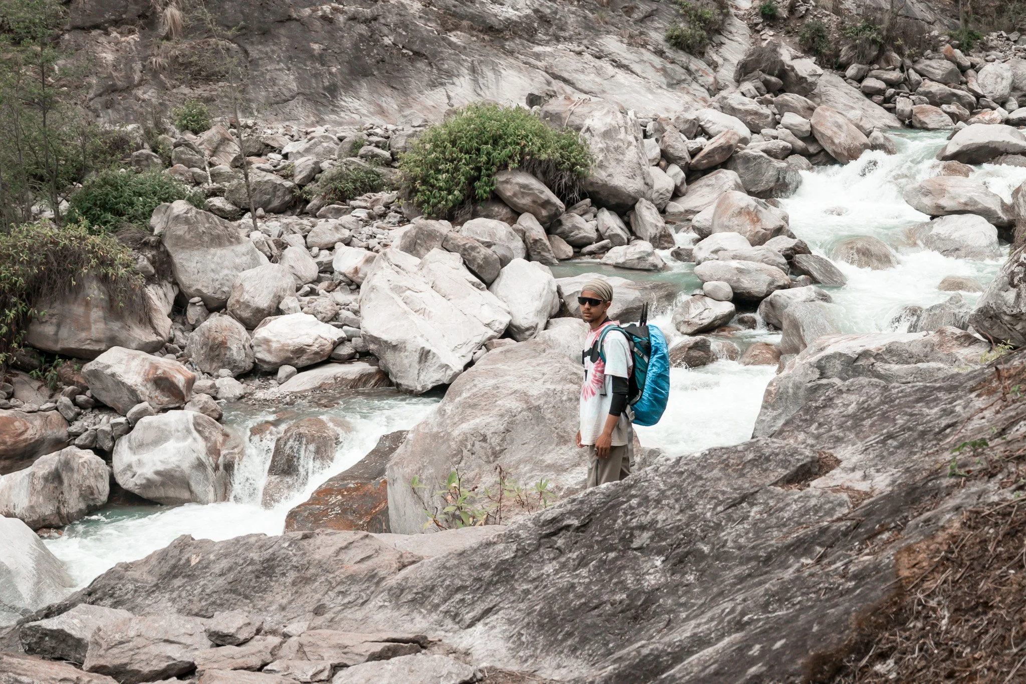 A man wearing sunglasses, a head covering, a long-sleeve shirt, and carrying a backpack, stands on a large rock in a mountain stream surrounded by rocks and sparse vegetation.