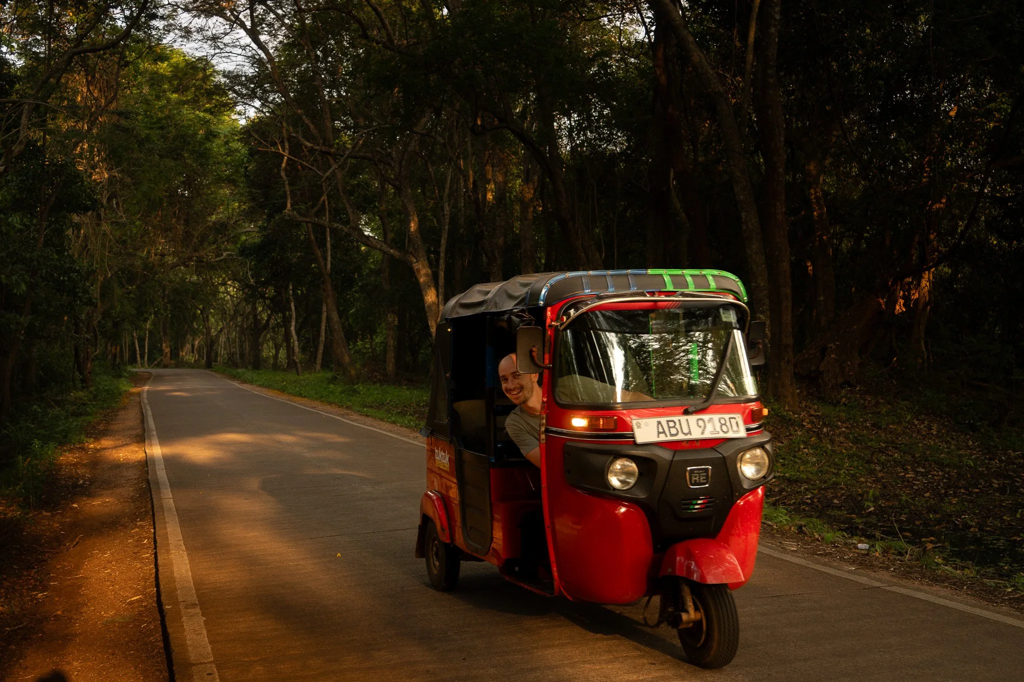 A red and black tuk-tuk driving on a narrow road through a wooded area.