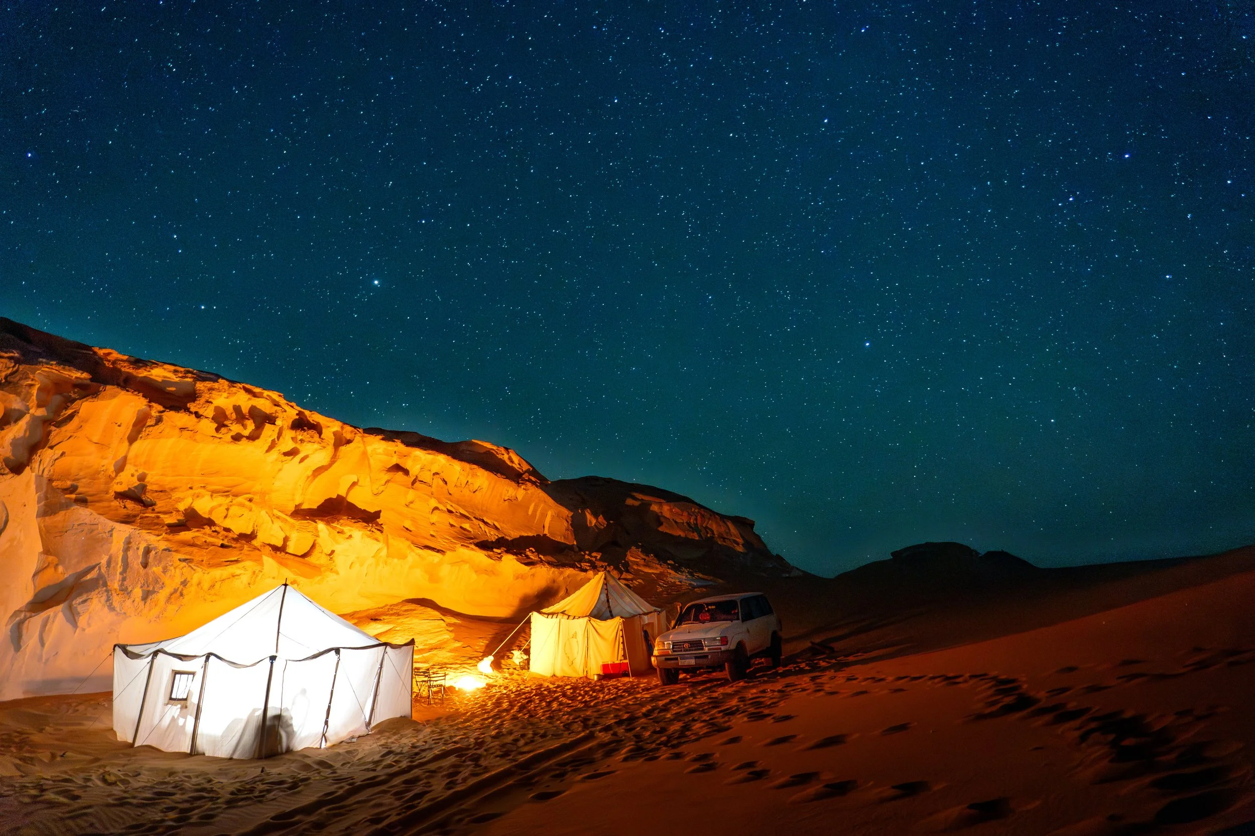 A desert campsite at night with illuminated tents, a vehicle, and a star-filled sky.
