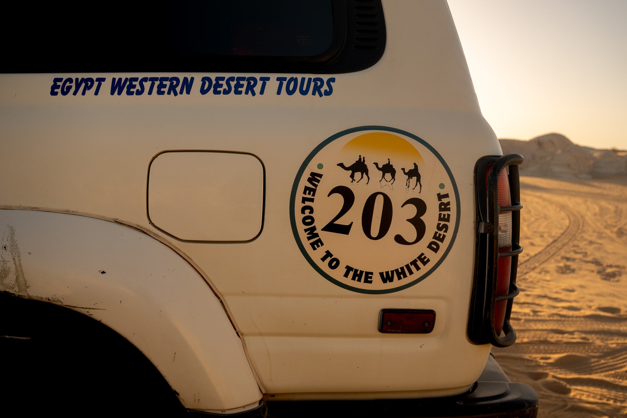 Back of a white desert tour vehicle with stickers, including 'Egypt Western Desert Tours' and a circular sticker that says 'Welcome to the White Desert 203' with illustrations of camels and a sunset, in a sandy desert landscape at sunset.
