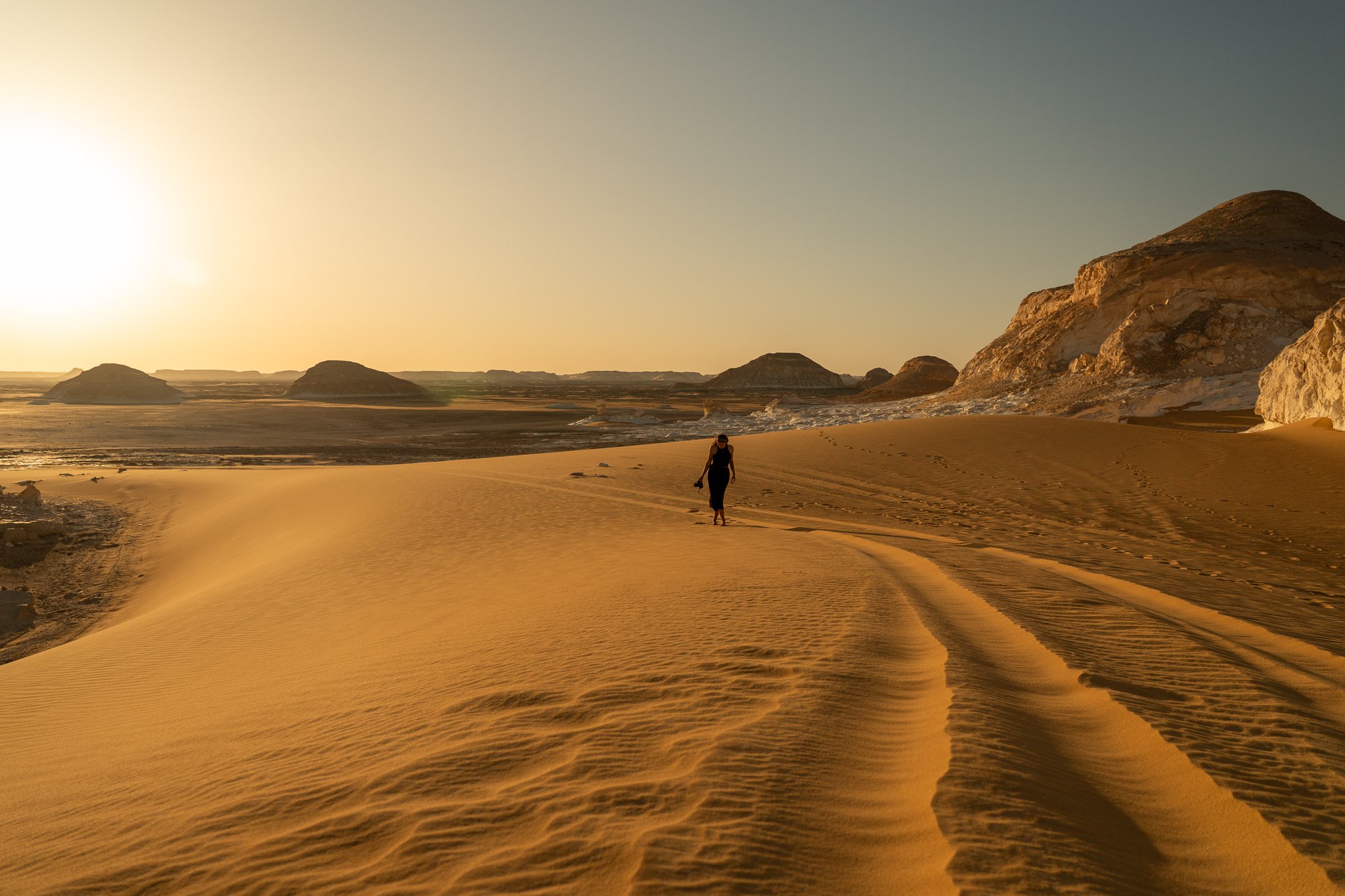 A person walking alone in a desert at sunset, surrounded by sand dunes and rocky formations.