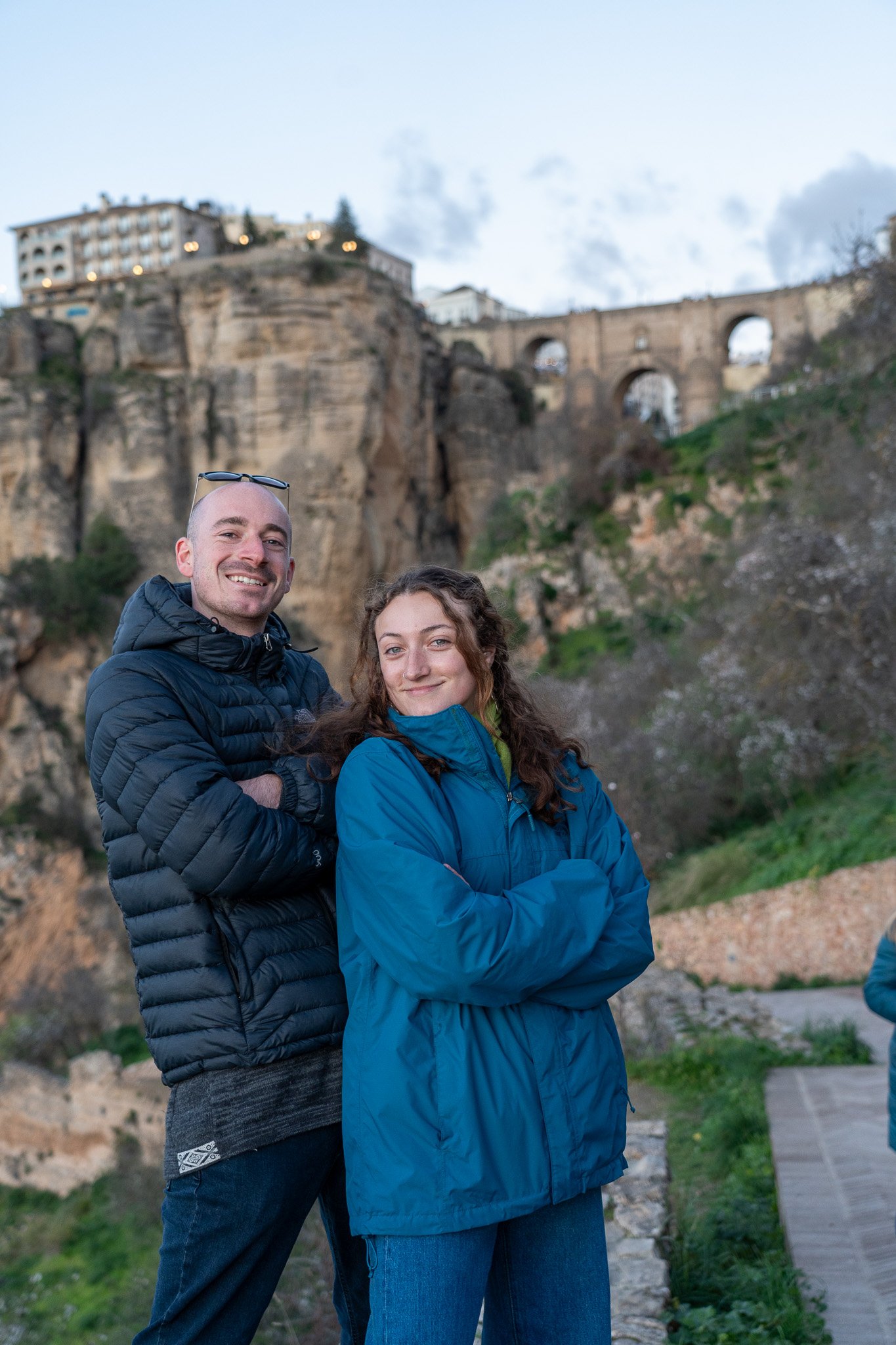 A young man and woman stand outdoors with a rocky hillside and historic stone arches in the background. They are smiling, wearing jackets, and have their arms crossed.