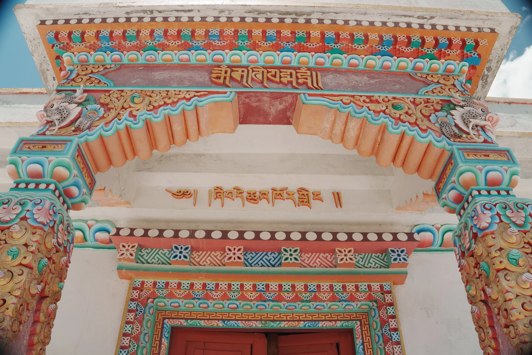 Colorful, ornate Tibetan or Himalayan temple entrance with decorated pillars, intricate carvings, and painted details, including dragons and traditional symbols.