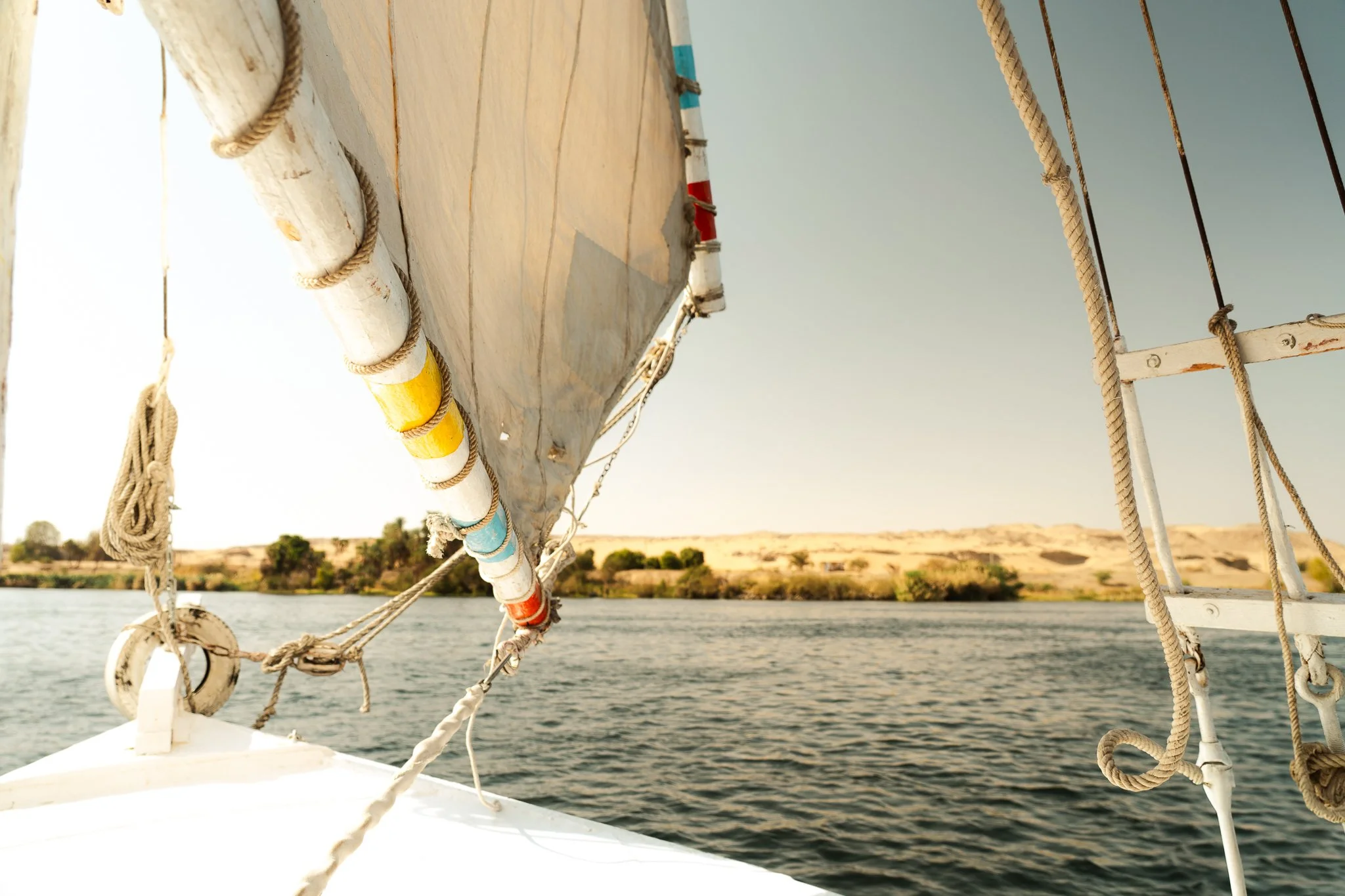 View from a boat looking at the water with a shoreline and hills in the distance, part of the boat's sail, and rigging ropes visible in the foreground.