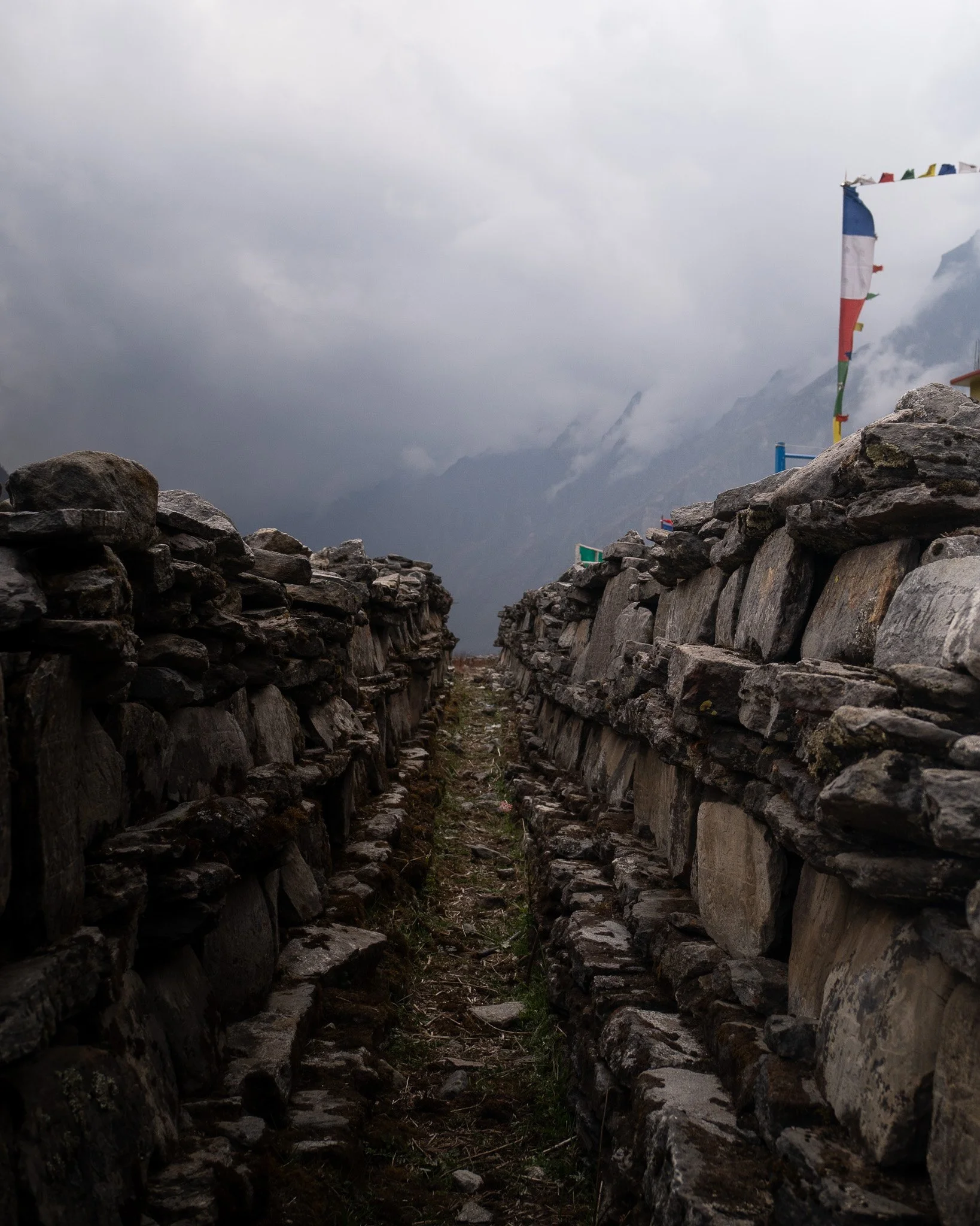 A narrow stone pathway between two stone walls, with mountain and cloudy sky in the background, and prayer flags on the right side.