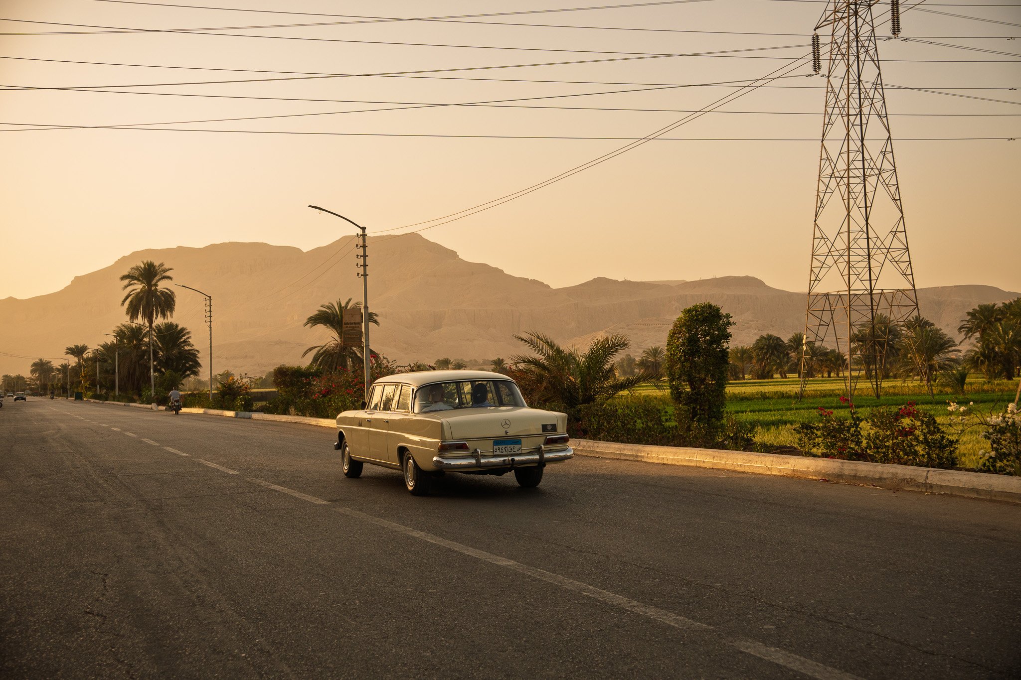 A vintage beige Mercedes-Benz car driving on a wide road with few cars and a motorcycle in the distance, lush green vegetation on the side, palm trees, mountainous landscape in the background, and a clear dusk sky filled with warm tones.