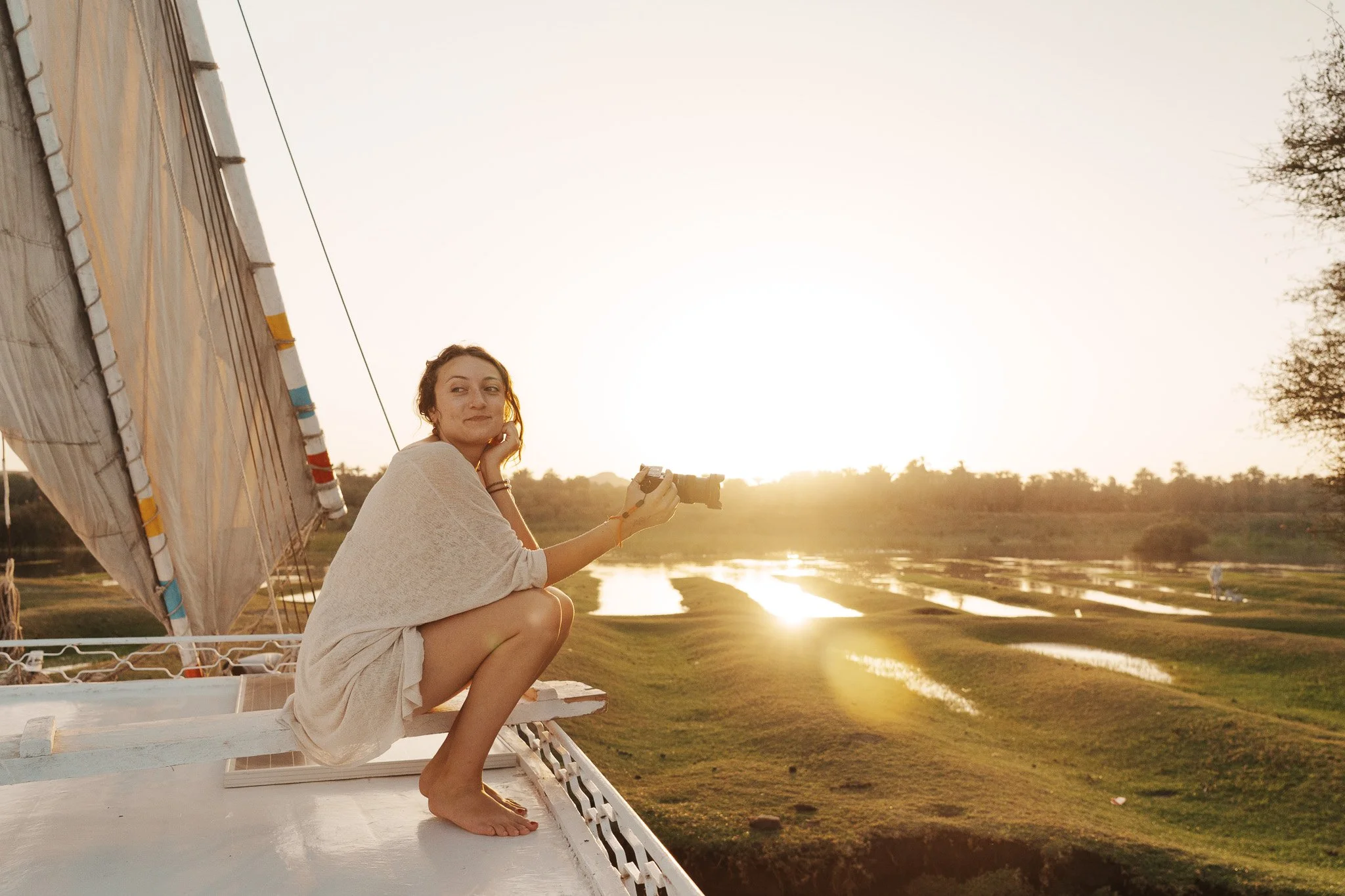 Woman sitting on boat, holding camera, during sunset over a river with fields and trees in the background.