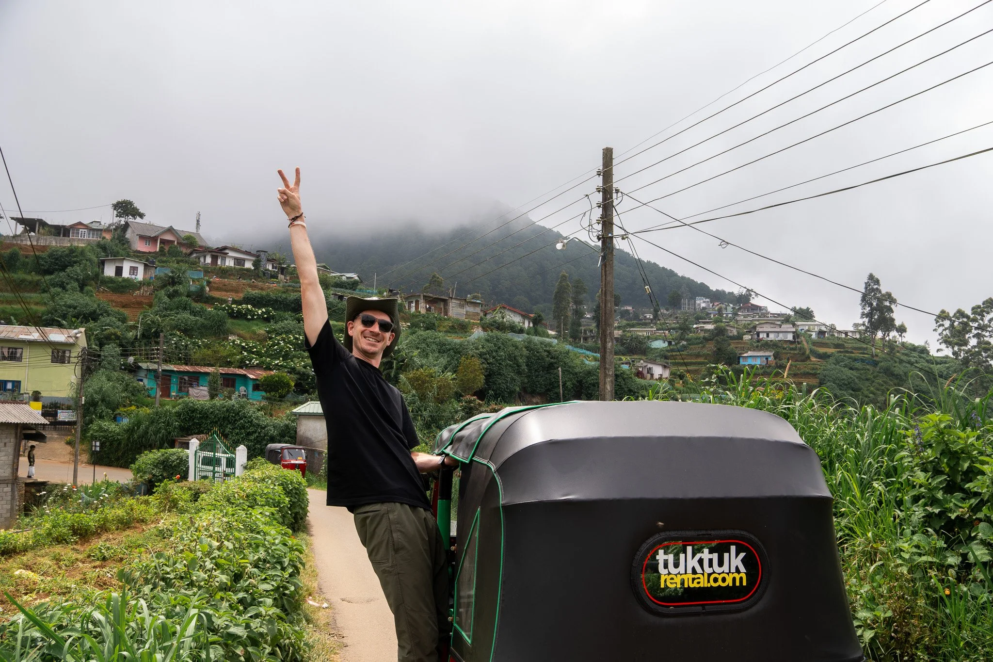 A man smiling and holding up a peace sign while riding in a tuk-tuk on a rural mountain road, with lush green plants, houses, and utility poles in the background.