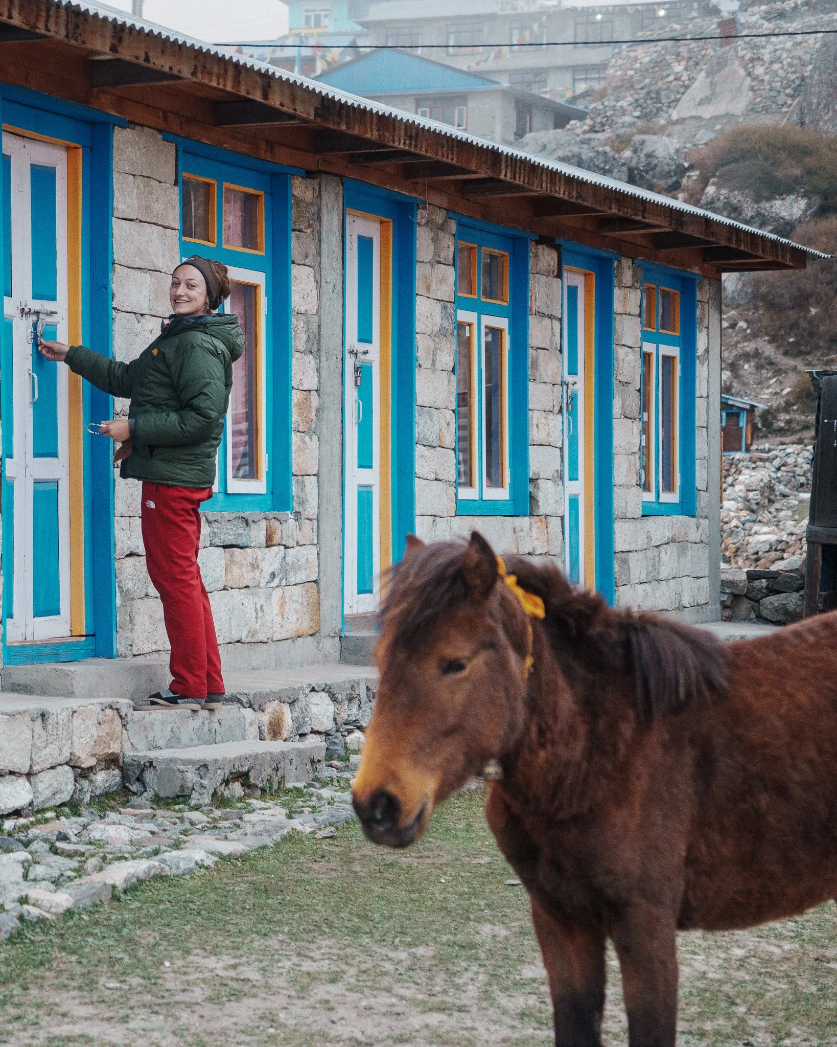 A woman with a green jacket and red pants standing on stone steps in front of a building with stone walls and blue-framed windows, smiling and unlocking the door. A brown horse with a yellow ribbon in its mane is in the foreground.