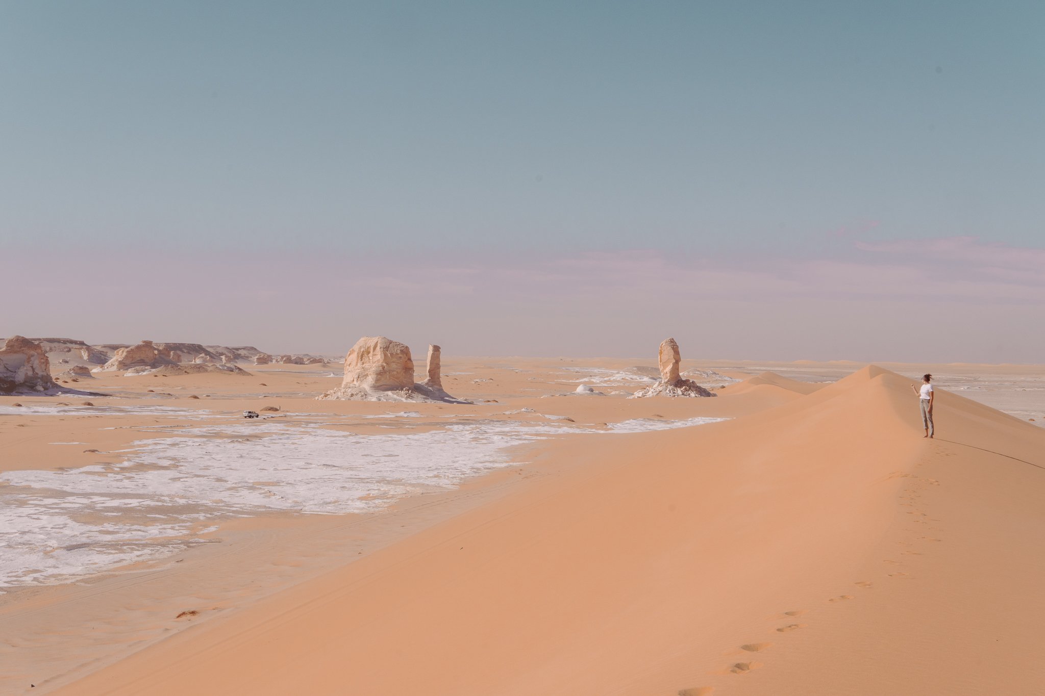A person stands on a sand dune in a desert with rock formations in the background and a clear blue sky.