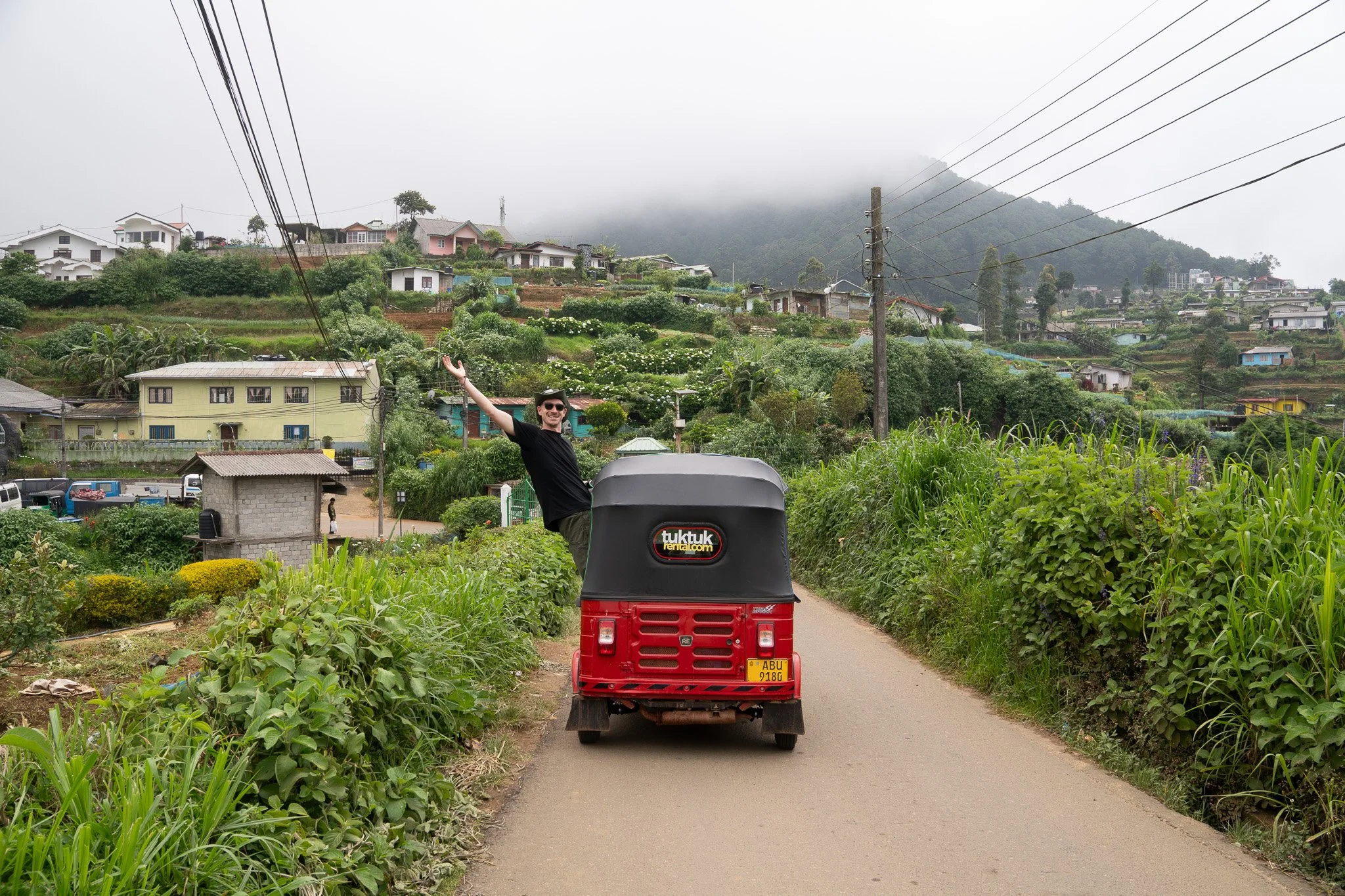 A man is standing in a red and black tuk-tuk with his arms raised, on a narrow rural road surrounded by green plants and hills, with houses and power lines visible in the background, and a foggy mountain in the distance.