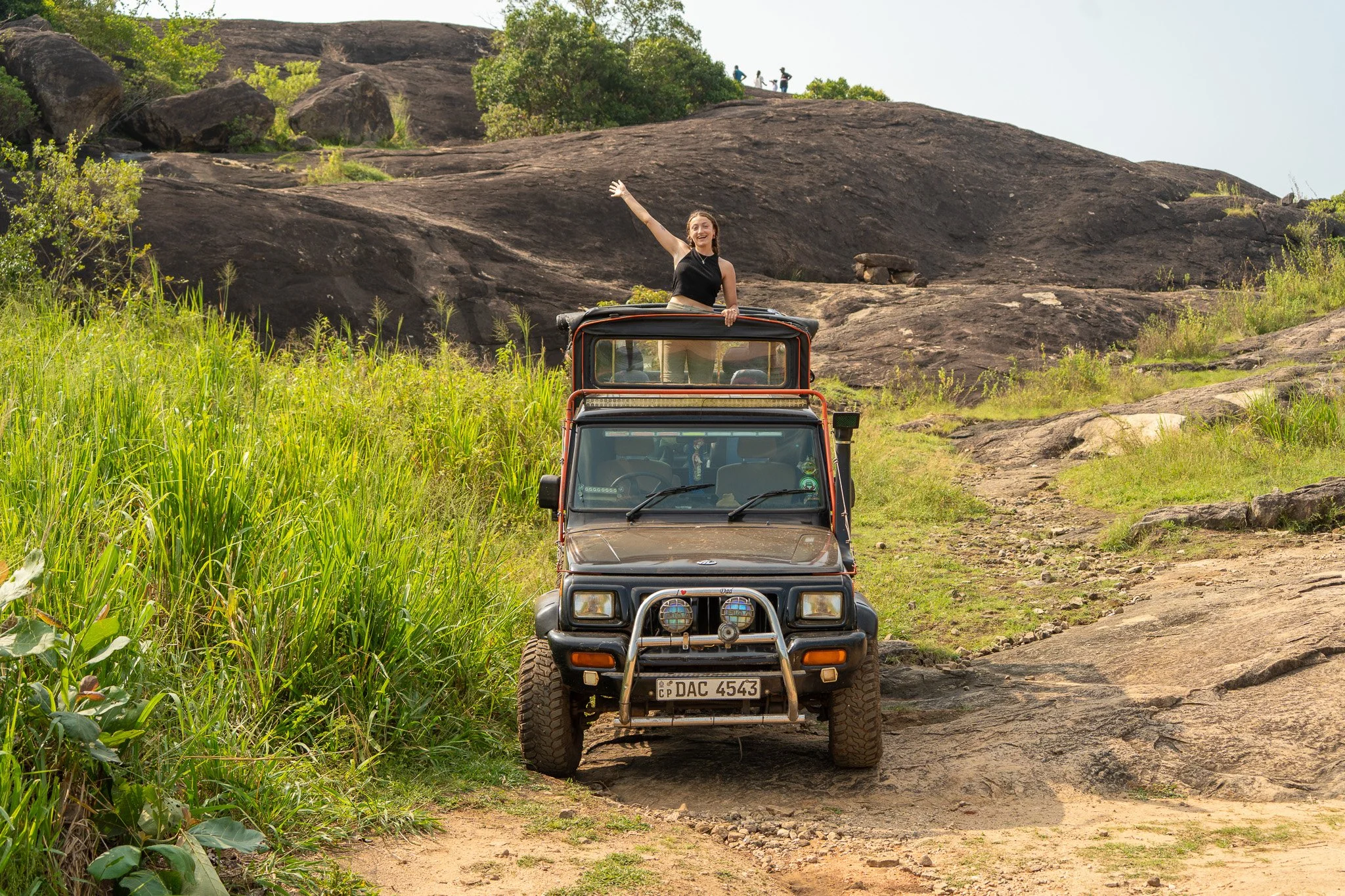 A woman is standing through the sunroof of a parked off-road vehicle on a rocky trail in a grassy hillside, waving and smiling with a scenic natural landscape of rocks, grass, and trees in the background.